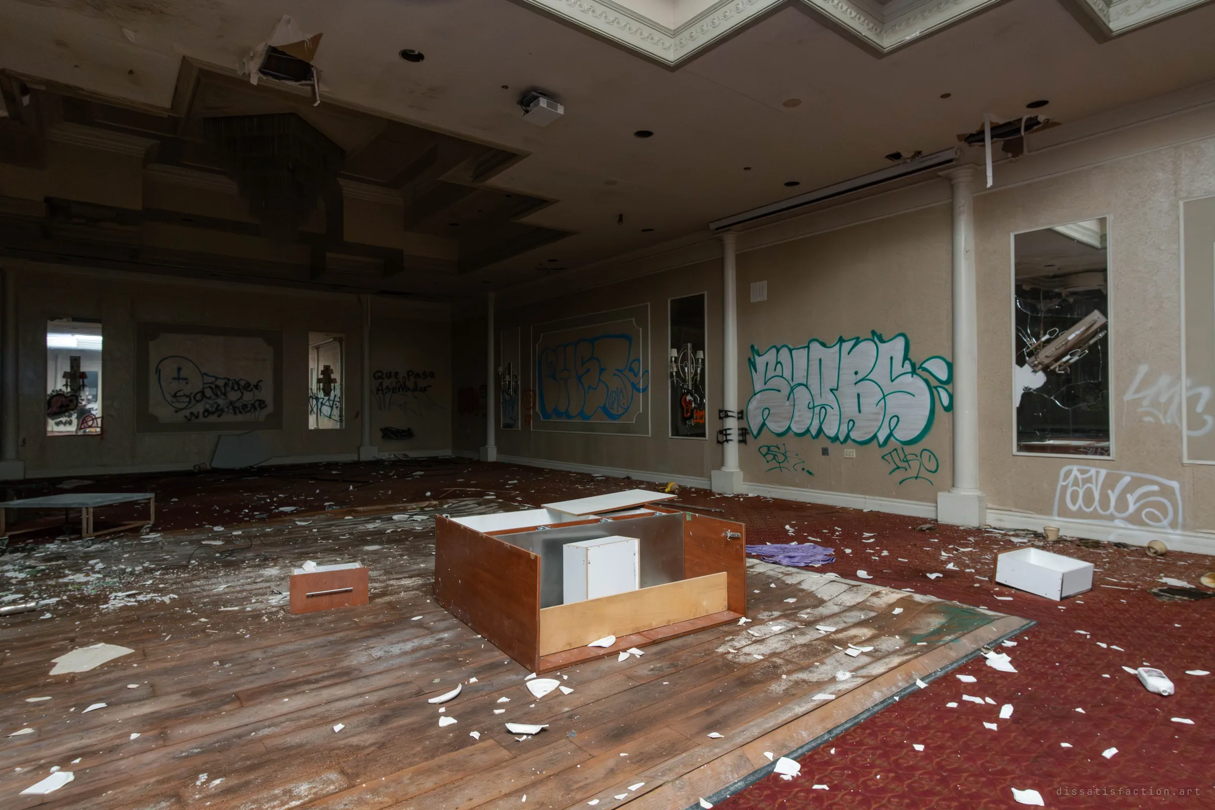 A dim room seen at an angle showing the wooden dance floor with an overturned cabinet in the middle, graffiti on the walls and cracked mirrors reflecting a chandelier.