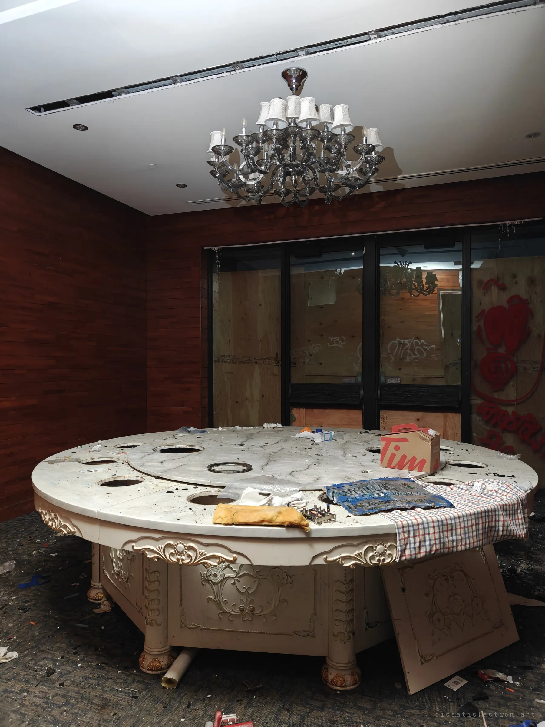 A large round marble banquet table with a lazy-Susan top sits under a grey chandelier. A coffee-chain box, a checked shirt and a hand-painted board lie on it, and the doors behind are boarded with plywood and red graffiti.