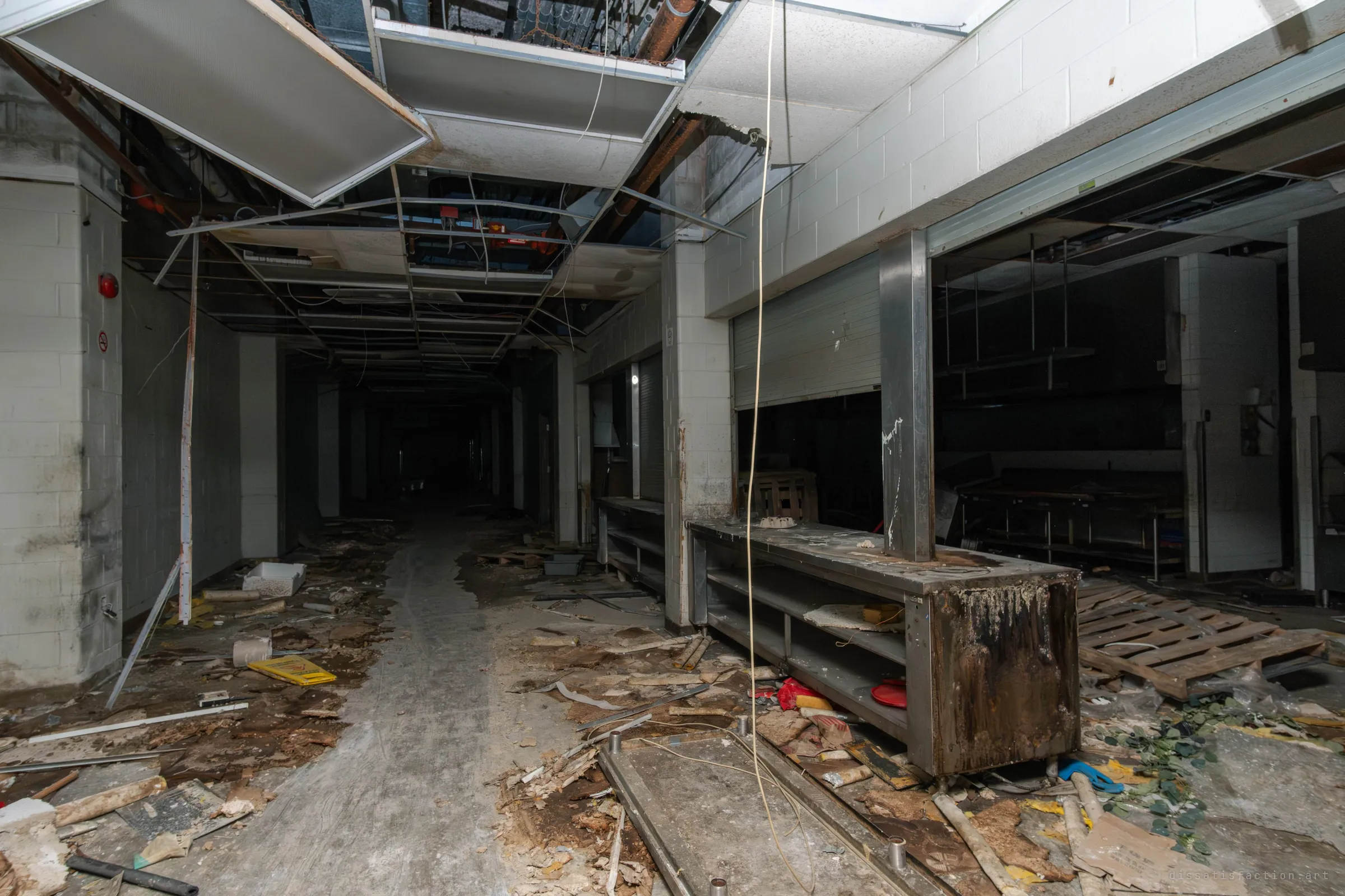 A dark, debris-strewn kitchen corridor with a collapsed ceiling, hanging wires and a rusted steel serving counter beside rolled-down shutters. The wreckage trails into black at the far end.