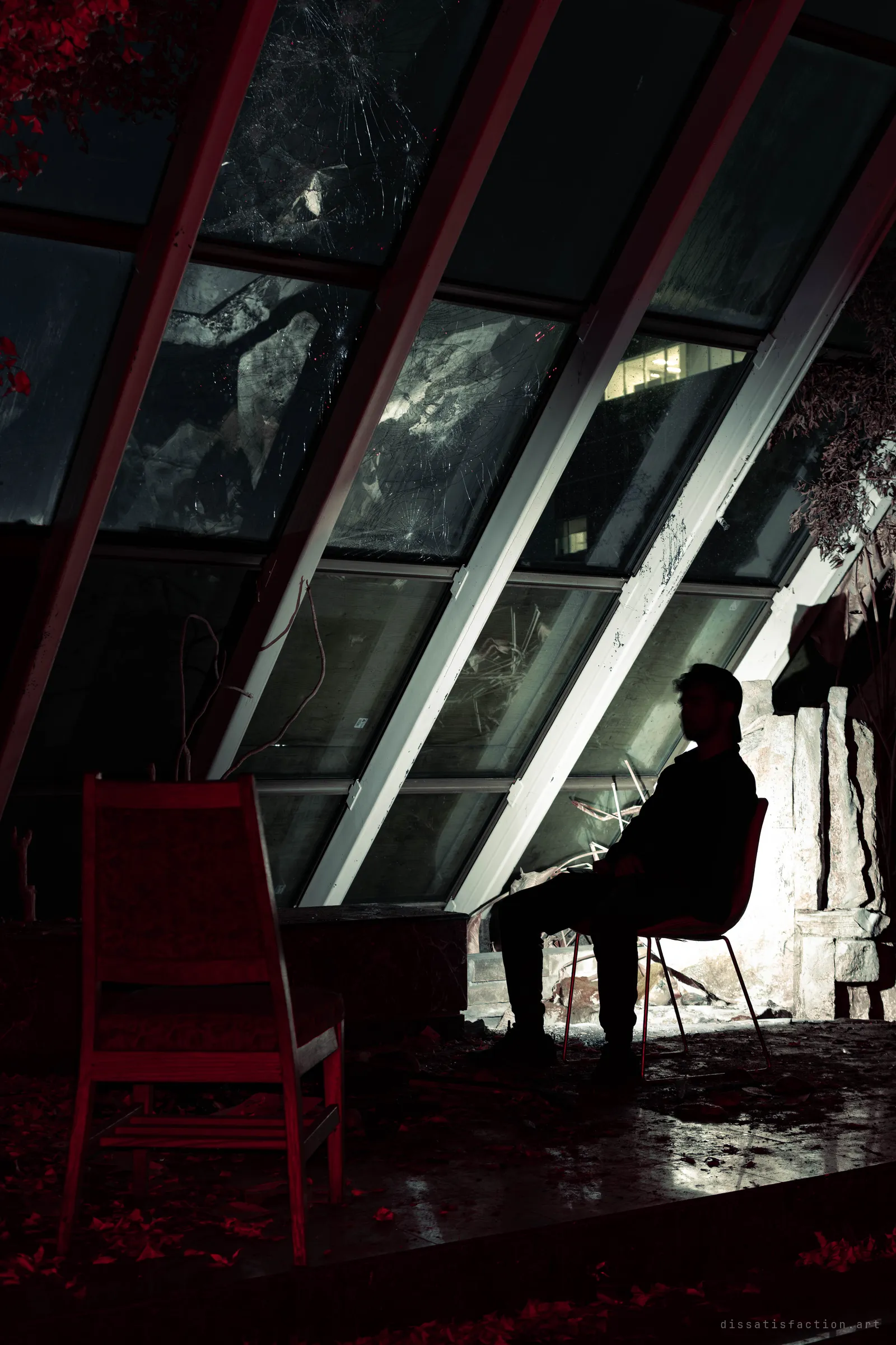 A figure in a cap sits in silhouette on a chair beside a spotlit stone feature, under a steeply slanted glass wall with shattered panes. Red-lit leaves and an empty chair fill the foreground.