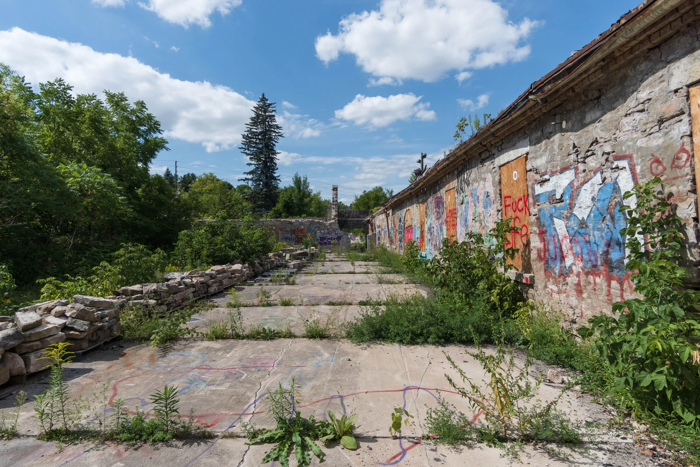 A cracked concrete yard runs between stacked salvaged stone and a long row of boarded, graffiti-tagged openings. Weeds push through every seam of the slab.