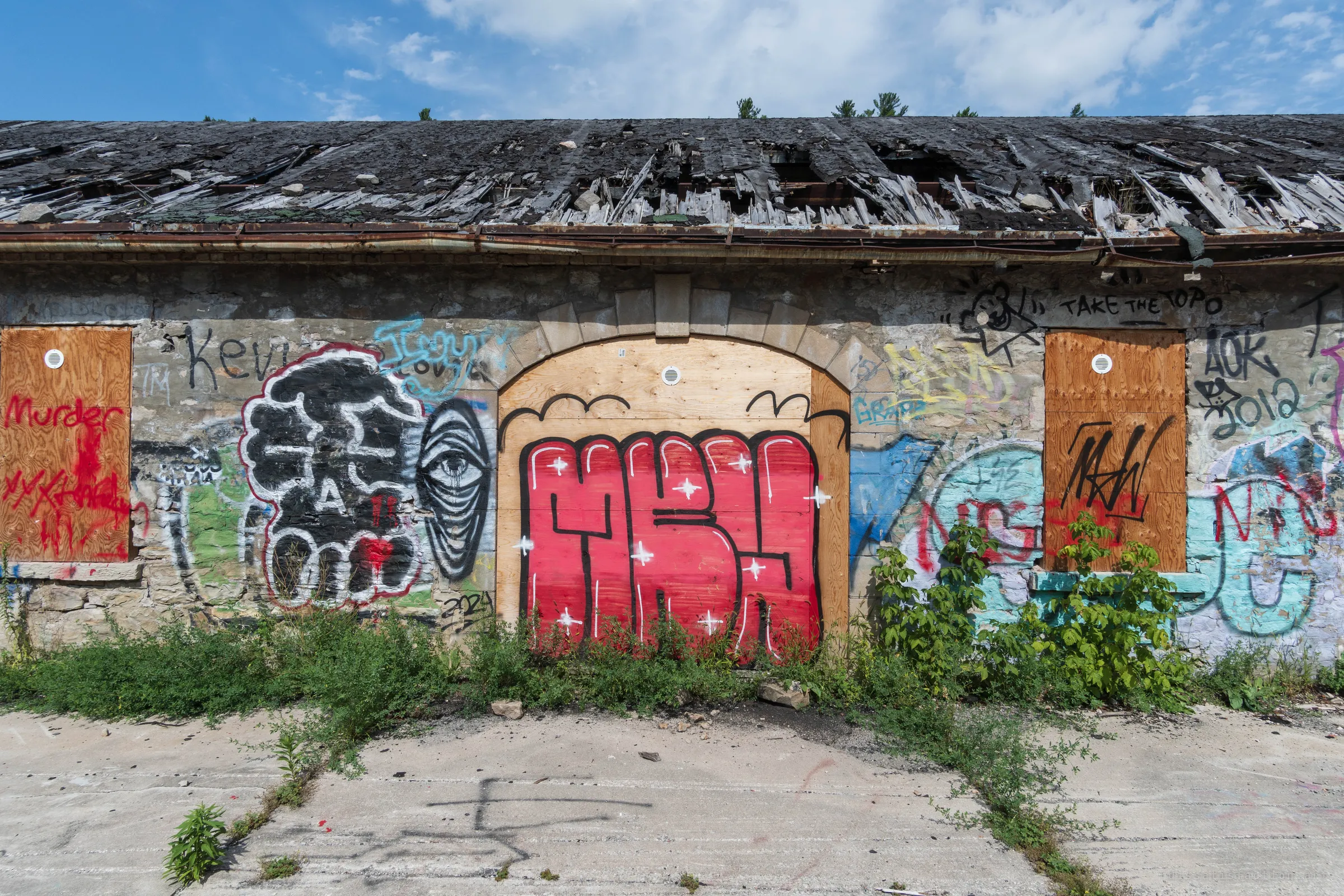 Head-on view of a stone shed with a collapsing plank roof, its arched doorway and windows boarded and layered in tags and throw-ups. The caving roof shows the building slowly giving way from the top down.