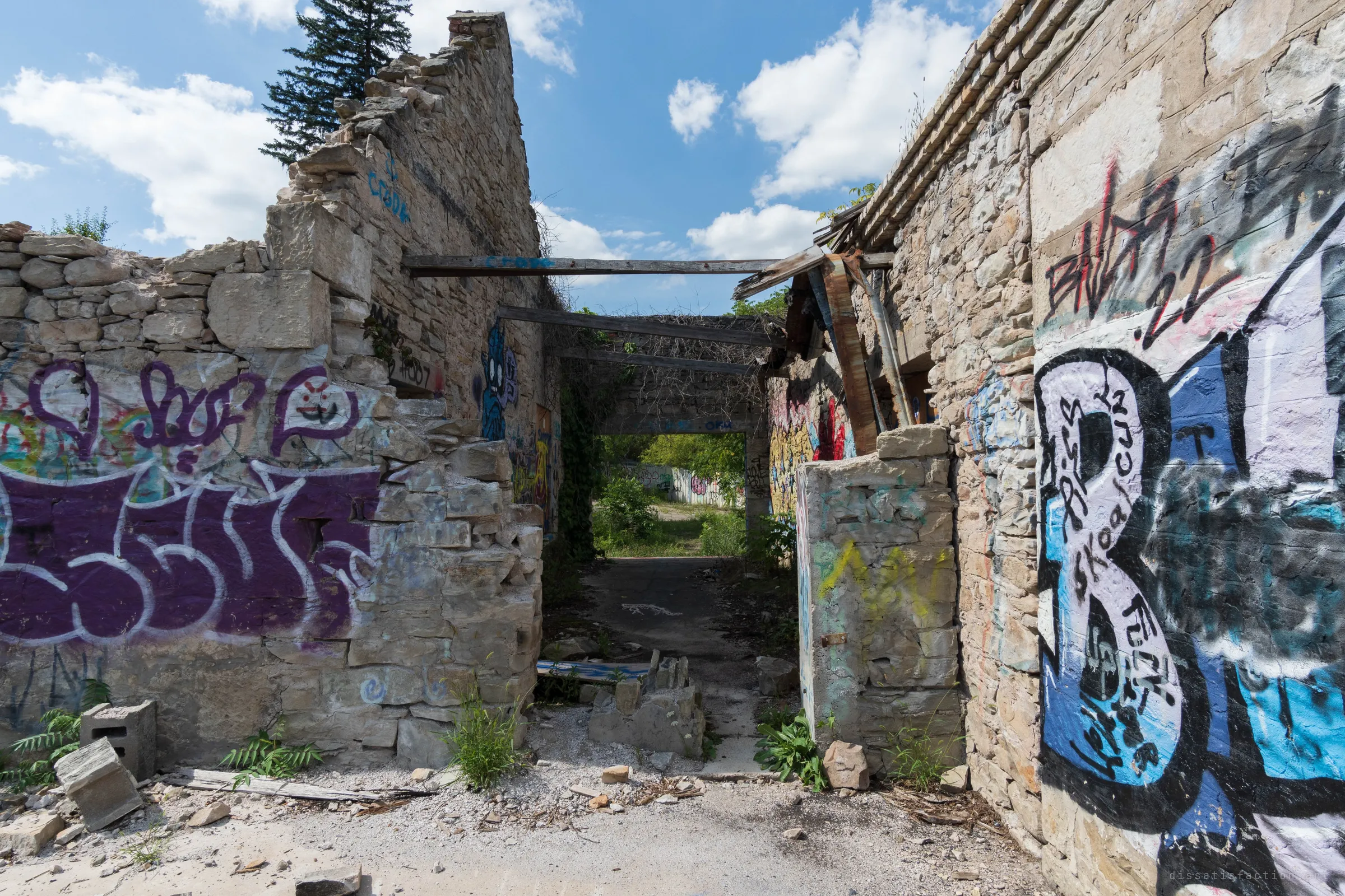 A narrow passage between crumbling rubble-stone walls, spanned by bare timber beams and open to the sky. Graffiti covers every surface and green light glows from the yard beyond.