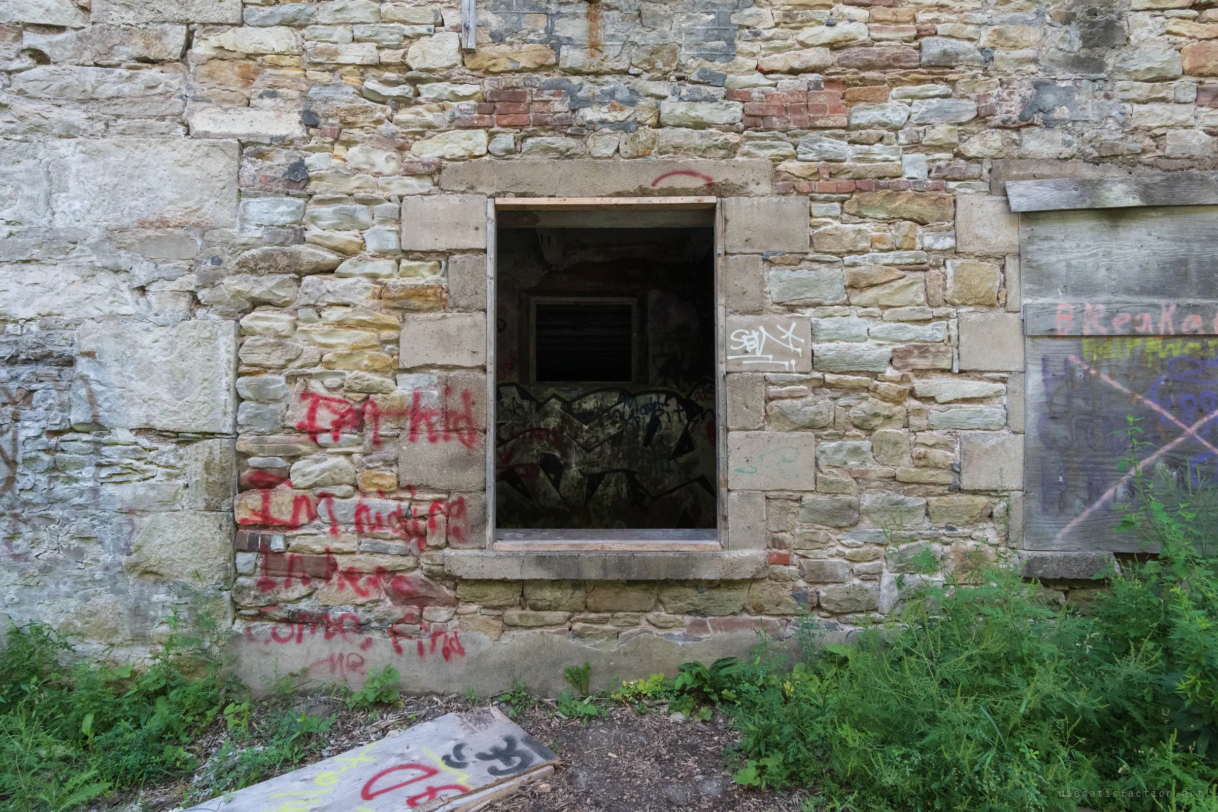 A single dark window opening set in a patched stone and brick wall, with a painted interior visible inside and a scrawled message running down the masonry beside it. The mix of stone and later brick repairs shows the building's long working life.