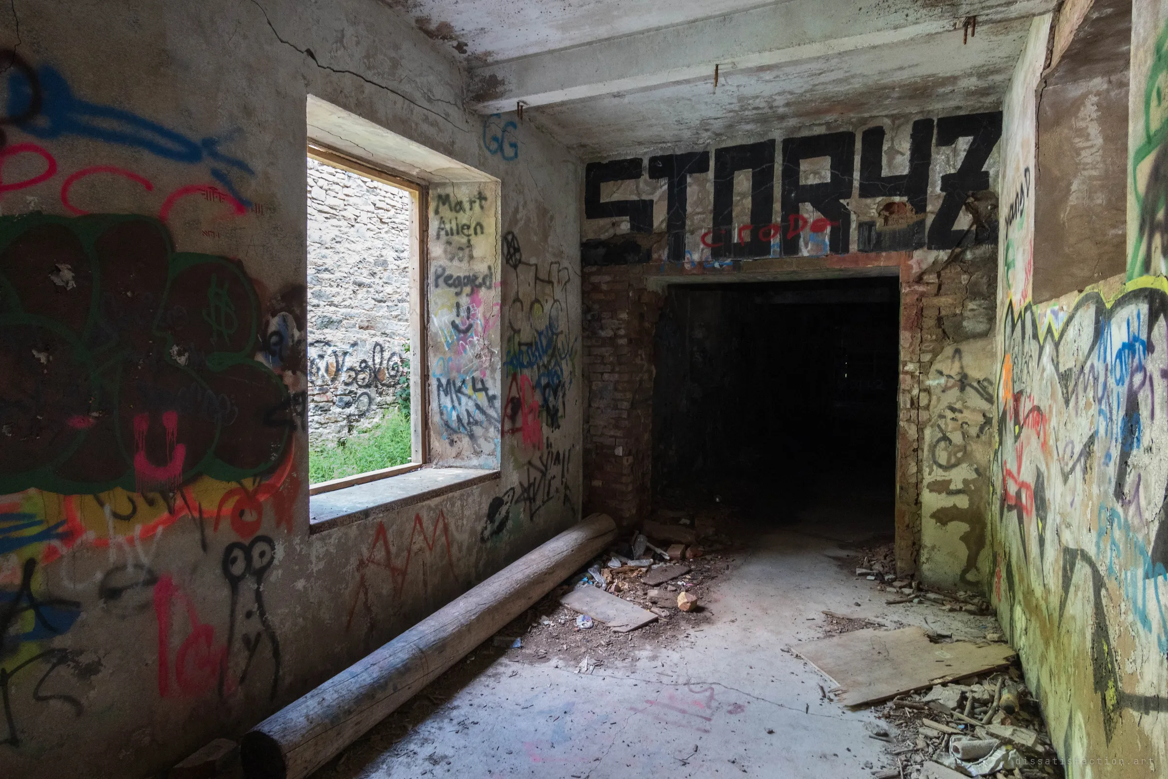 A graffiti-covered interior room with a window onto a stone wall outside and a dark doorway under a big black tag. A log and fallen boards lie across the littered floor.