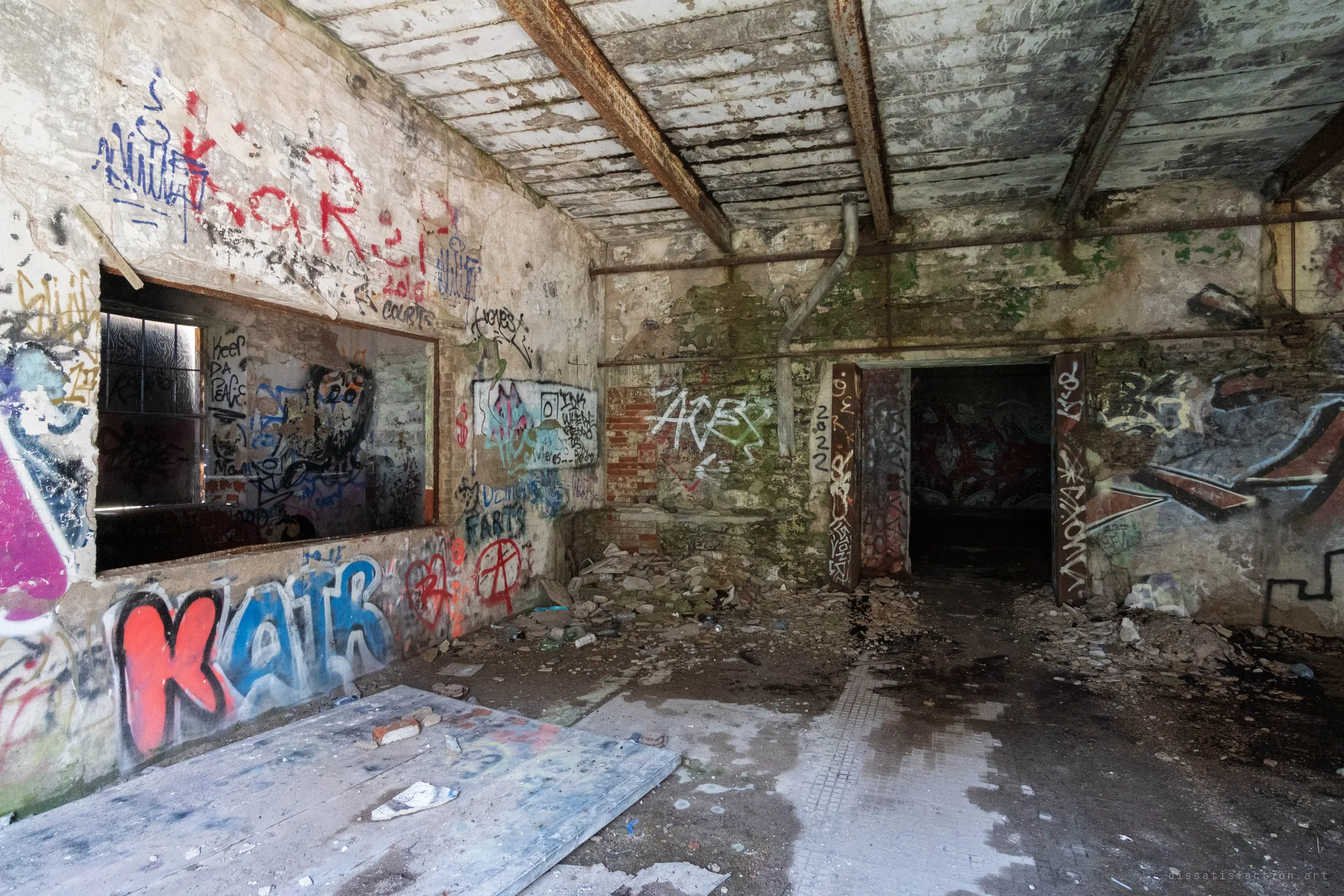 A room with rusted steel ceiling beams, peeling plaster and walls crowded with tags, opening through a dark doorway into a further painted chamber. Debris and puddles cover the tiled floor.