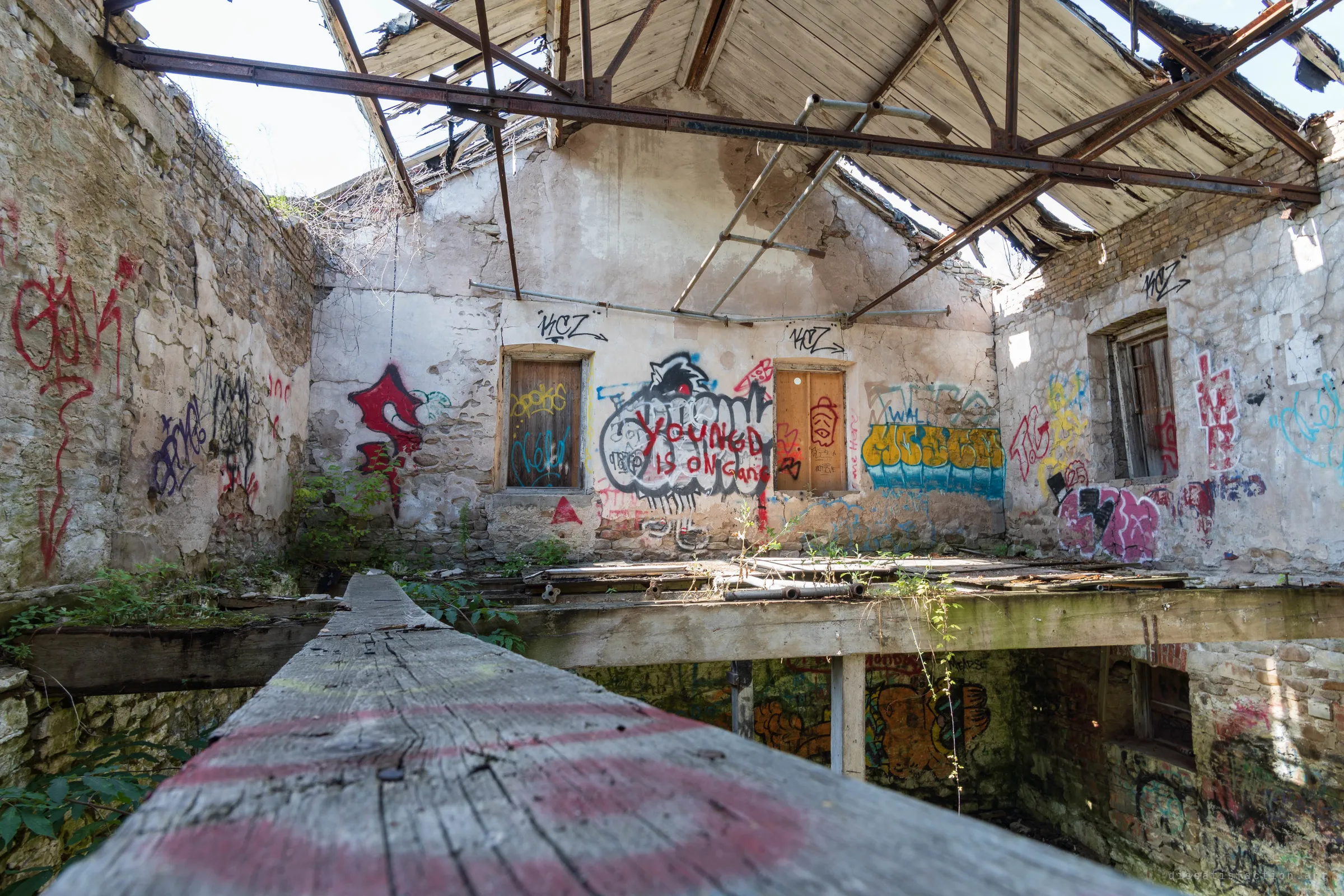 Looking along a paint-streaked timber beam into a roofless stone room, its gable wall covered in graffiti around two boarded windows. Rusted steel trusses and a sagging plank ceiling hang open to the sky, with a flooded lower level visible below the beam.