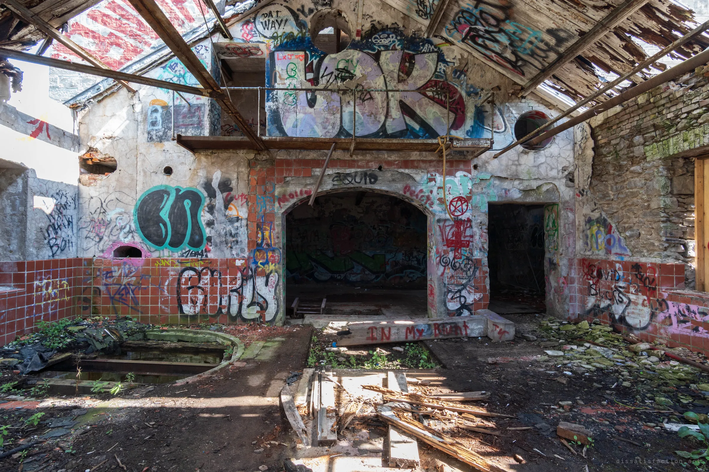 A roofless tiled hall packed with layered graffiti, with an arched opening and a steel catwalk under the collapsed roof. Debris, a sunken rim pit and scattered timber cover the floor in patches of sunlight.
