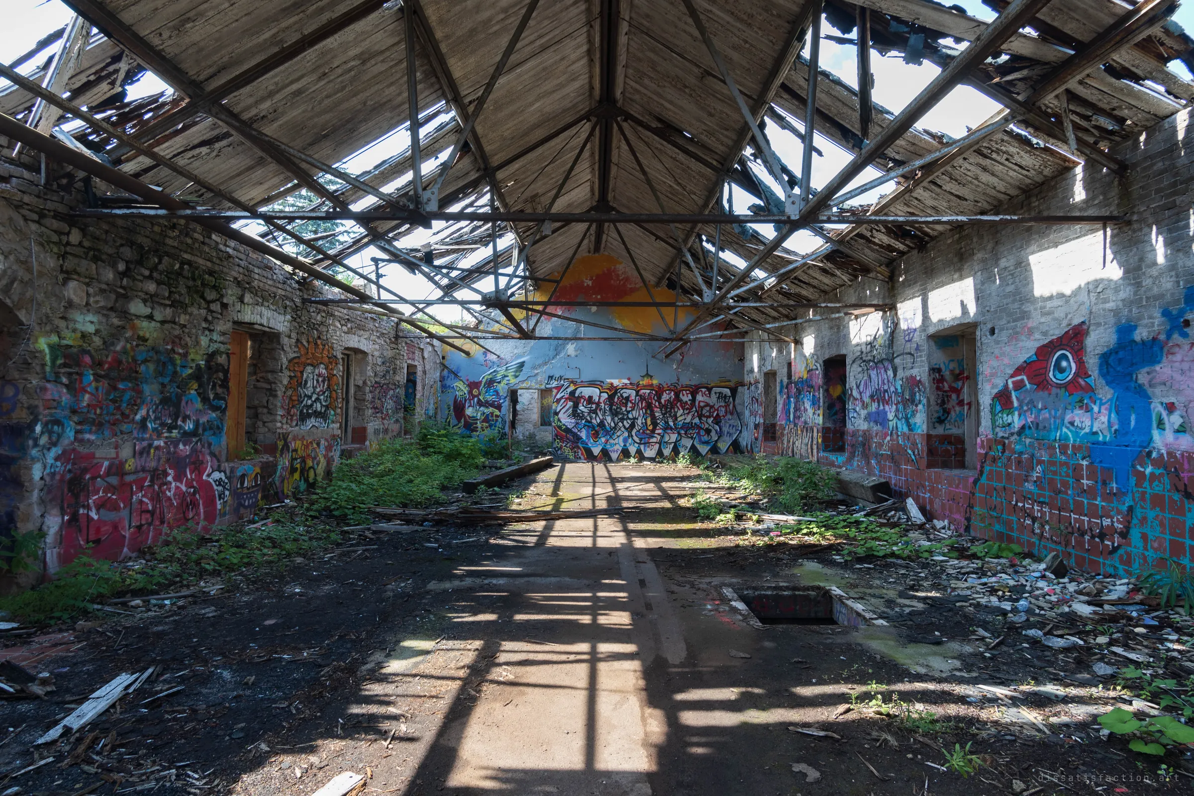 A long industrial hall with stone and brick walls covered in graffiti, open to the sky through a skeletal steel roof truss. Sunlight throws the truss shadows across an overgrown floor toward a mural on the far wall.