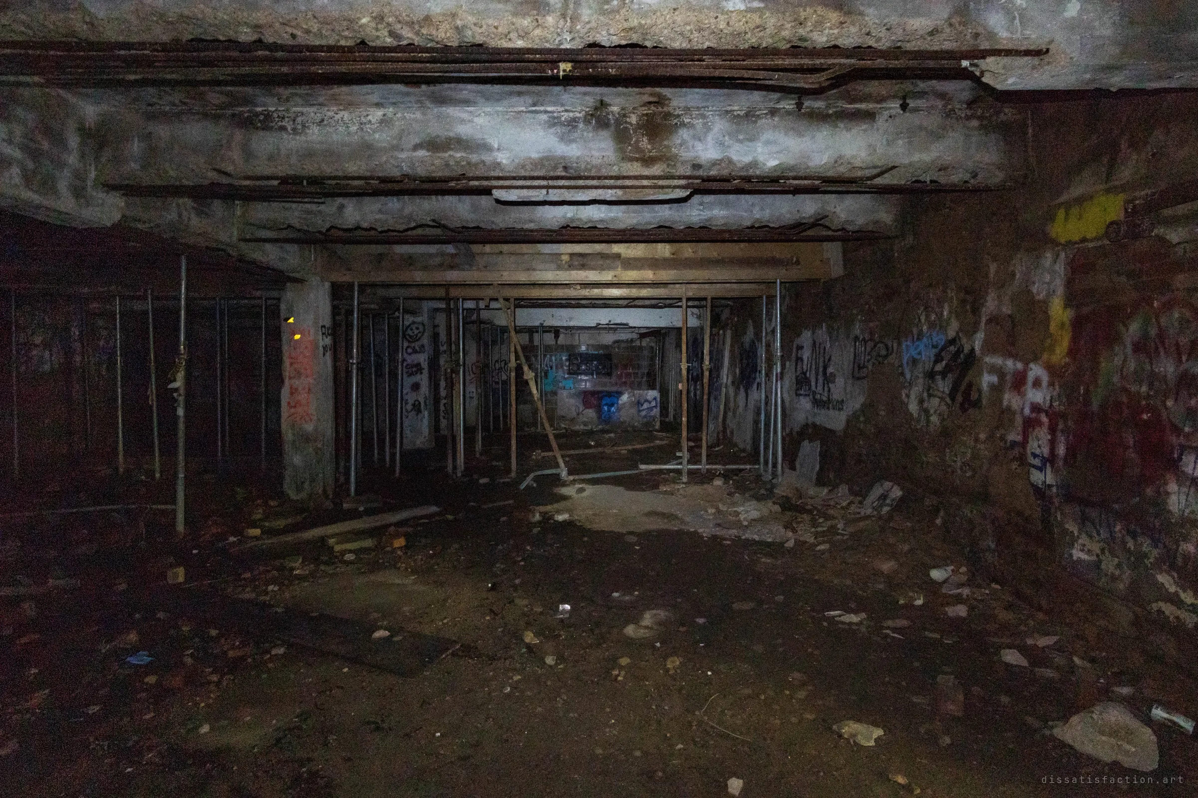 A dark basement level lit by flashlight, its low concrete ceiling held up by rows of steel shoring props. Rusted pipes run overhead and faint graffiti marks the walls beyond the debris-strewn floor.