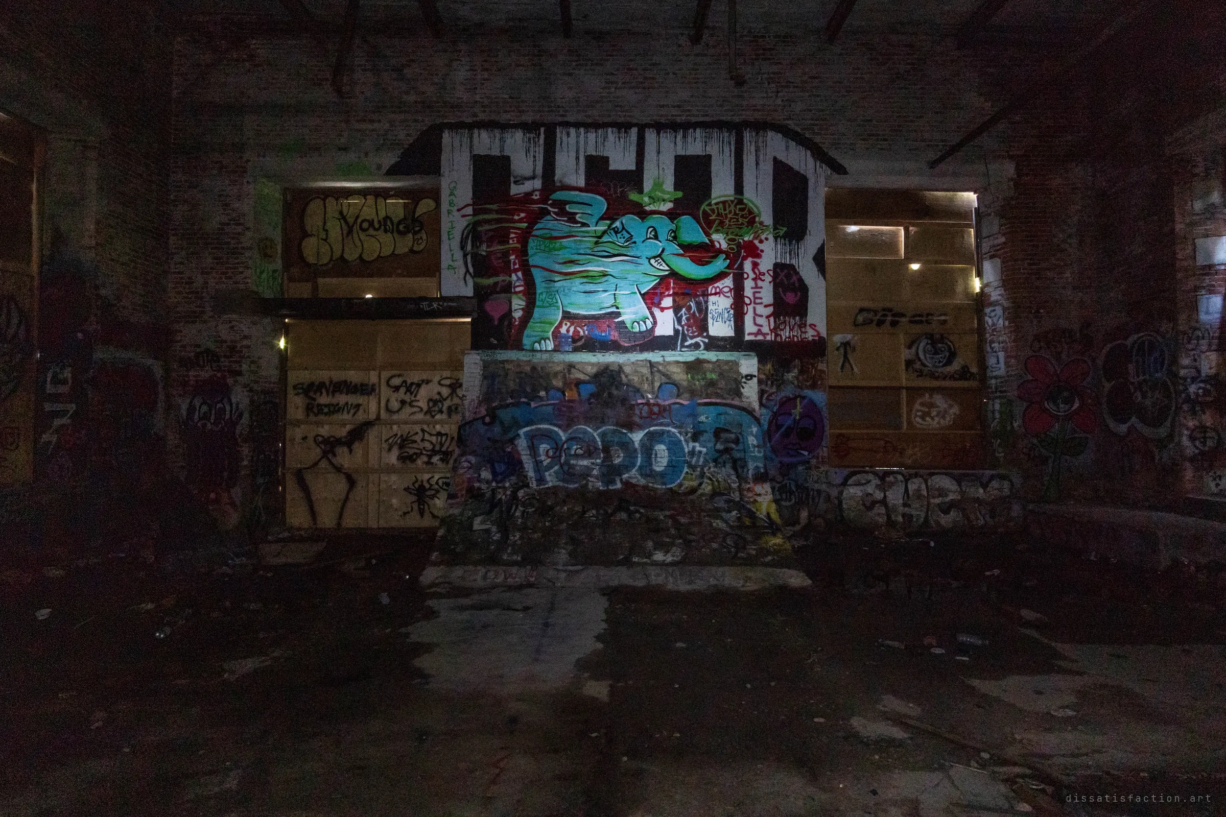 Head-on view of a dark wall where a flashlight picks out a cyan cartoon elephant mural above a graffiti-covered ramp. Boarded window bays with thin light leaking around their edges flank it on both sides.