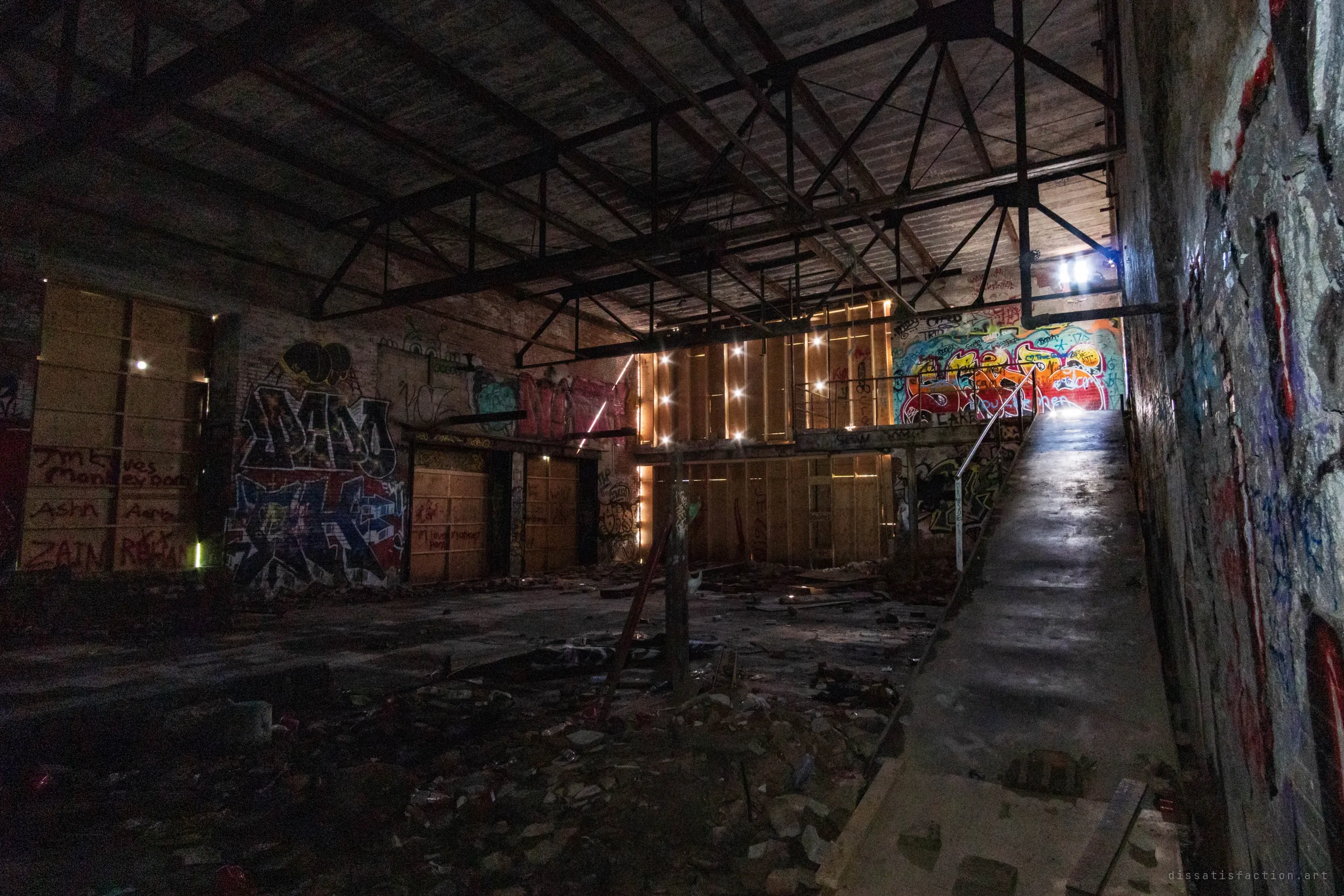 A cavernous dark hall under a steel roof truss, with a concrete ramp climbing along the right wall toward a bright graffiti-covered mezzanine. Light leaks through the boarded window bays in small points, like stars.