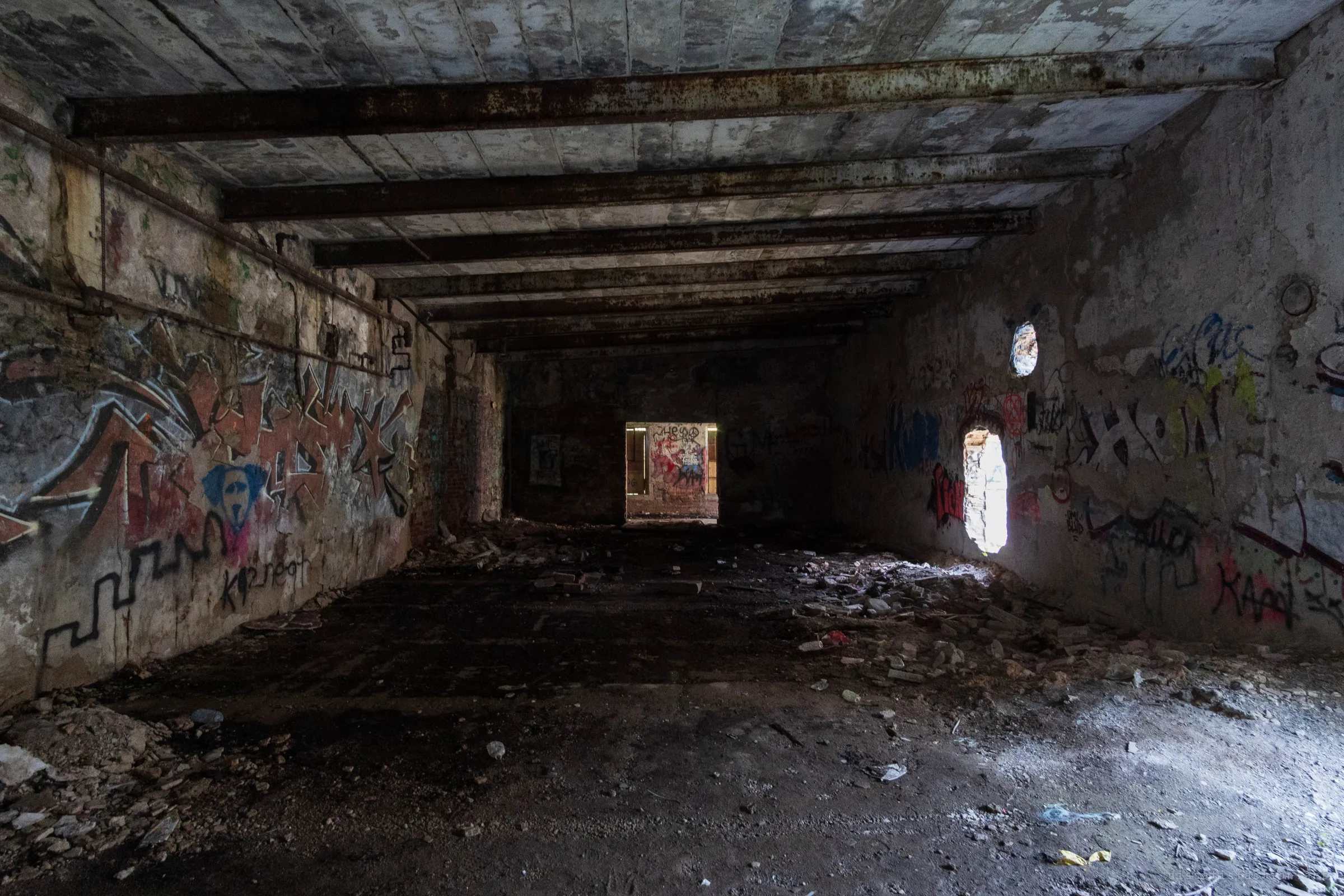 A long, low concrete room with rusted steel beams, its walls tagged with faded graffiti. Daylight falls through a narrow rounded opening onto a rubble floor, and a lit doorway glows at the far end.
