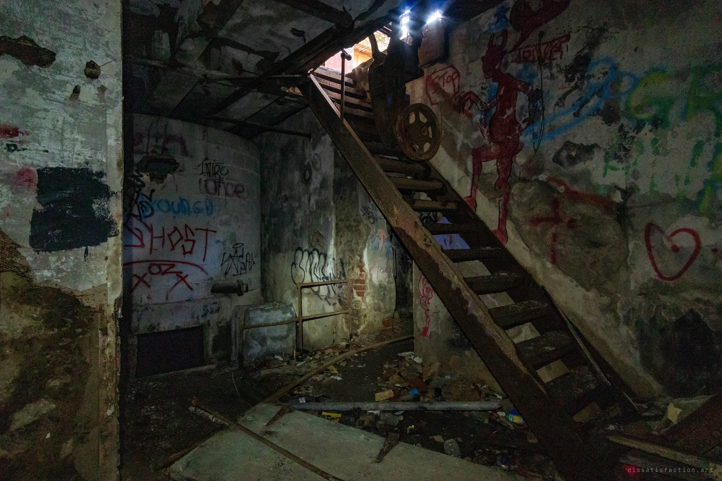 A rusted wooden staircase with an old wheeled machine climbs out of a graffiti-covered basement, torchlight catching a painted red devil on the wall. It shows how deep and layered the lower levels of the mill are.