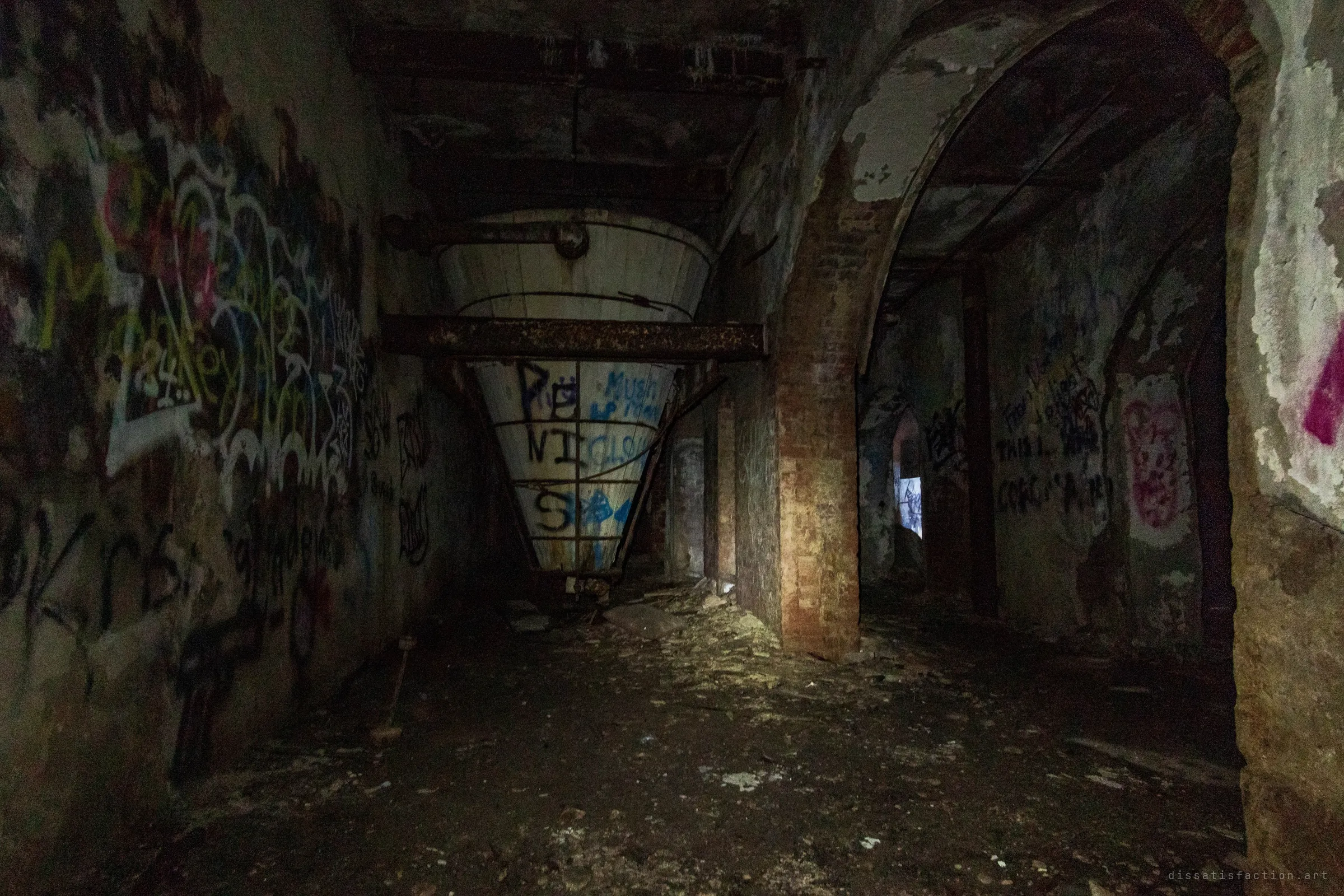 A huge inverted cone hopper hangs from steel beams in a dark brick-arched basement, its white sides tagged with graffiti. The hopper is one of the few pieces of process equipment still in place.
