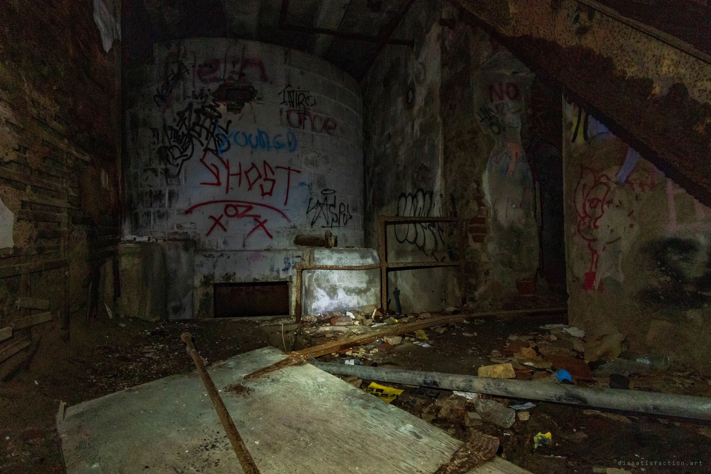 A closer look at the round graffiti-covered tank under the stairs, with planks, a fallen pipe and rubble strewn across the floor. The same basement corner as the staircase shot, seen head-on.