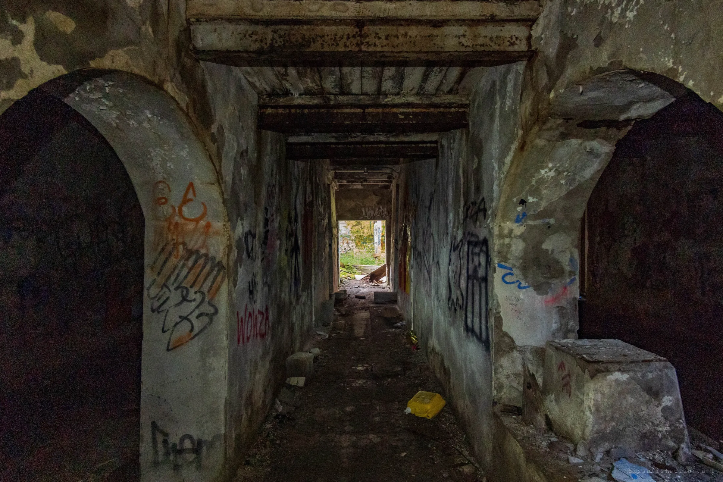 A narrow graffiti-lined passage runs between two brick arches toward a bright doorway full of weeds and daylight. It frames the way out from the dark basement.