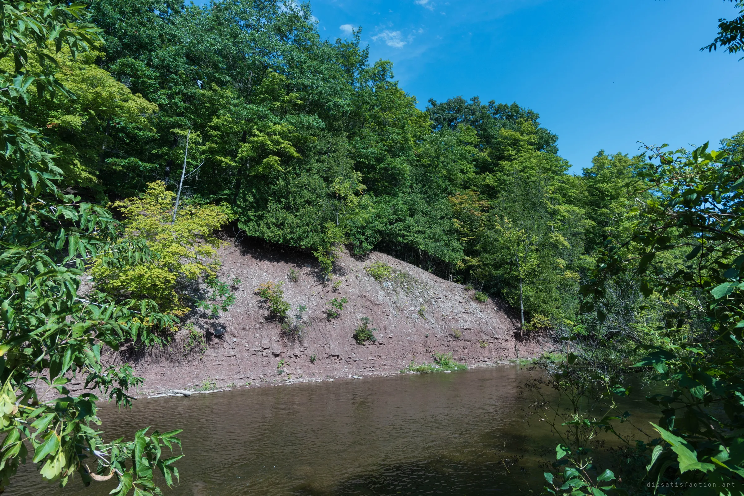 A calm brown river curves past a steep red shale bank under thick summer forest. The riverside setting explains why a mill was built here.