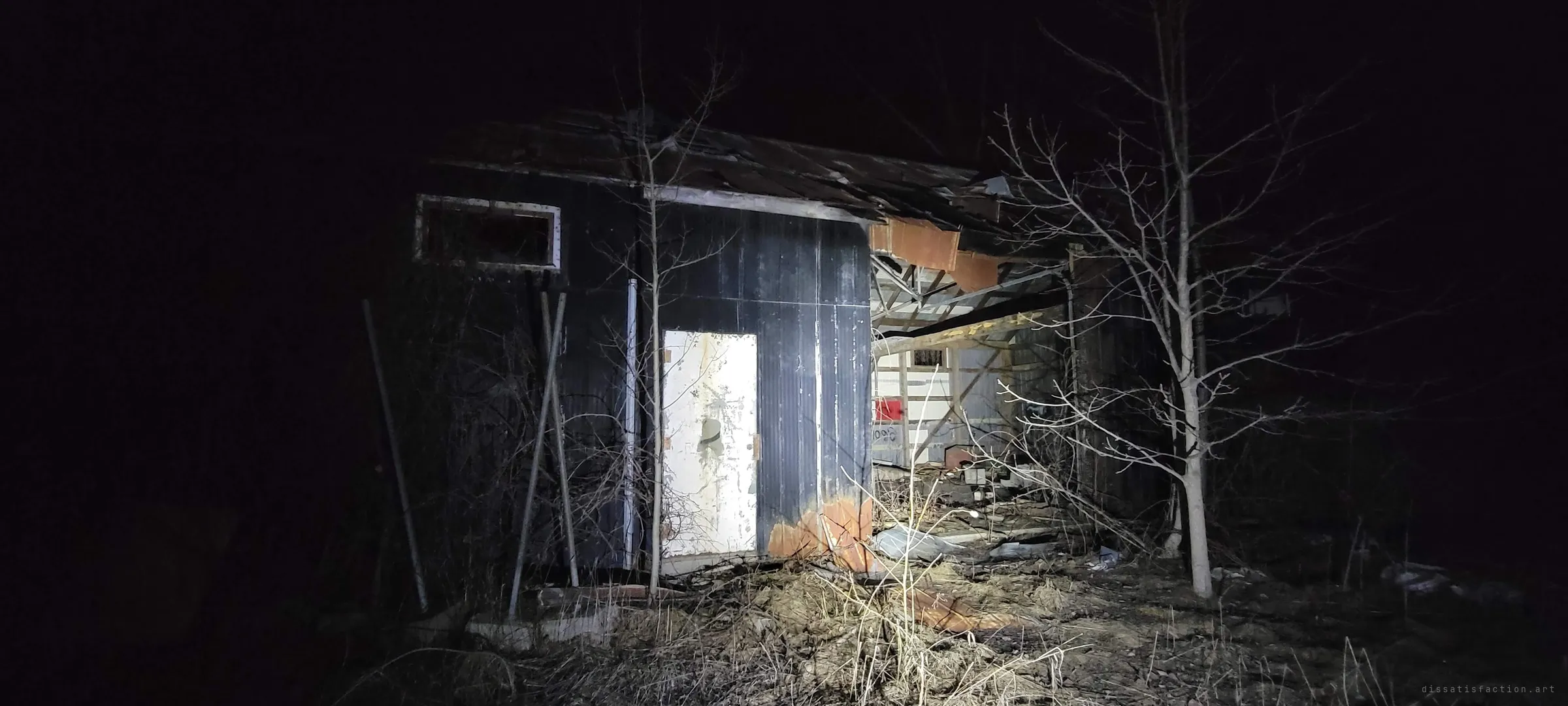 A dark-clad shed with a peeled white door and a caved-in roof, caught in flashlight at night among bare saplings and dead grass. The collapse opens the side of the building to the junk inside.