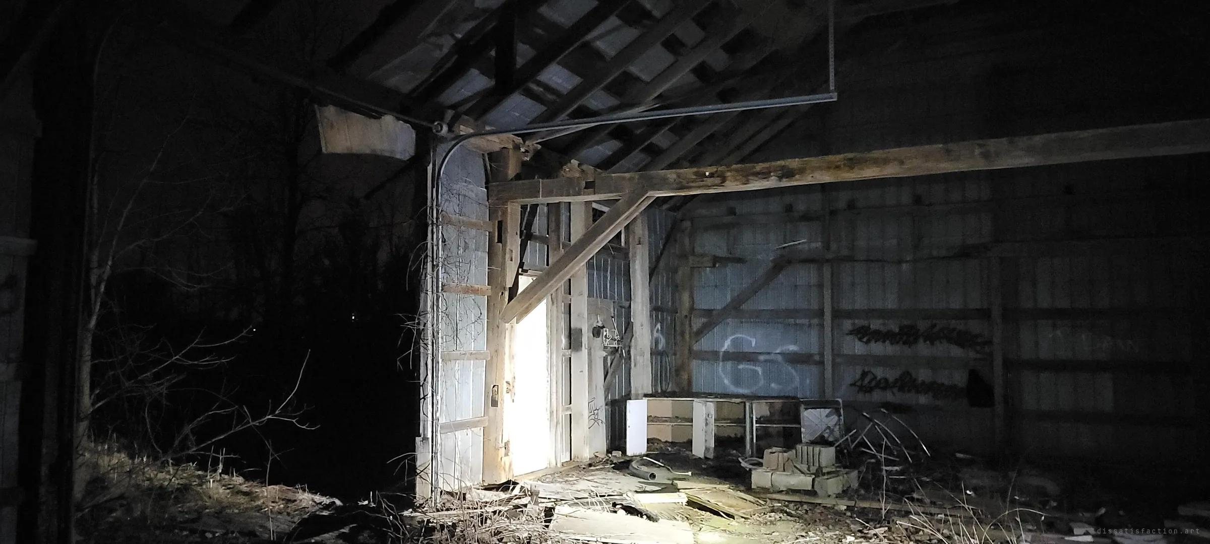 Inside a timber-framed barn, a garage door track hangs over a doorway lit white by a torch, with graffiti tags on the metal walls and debris on the floor. Heavy old beams still hold the roof over the mess.