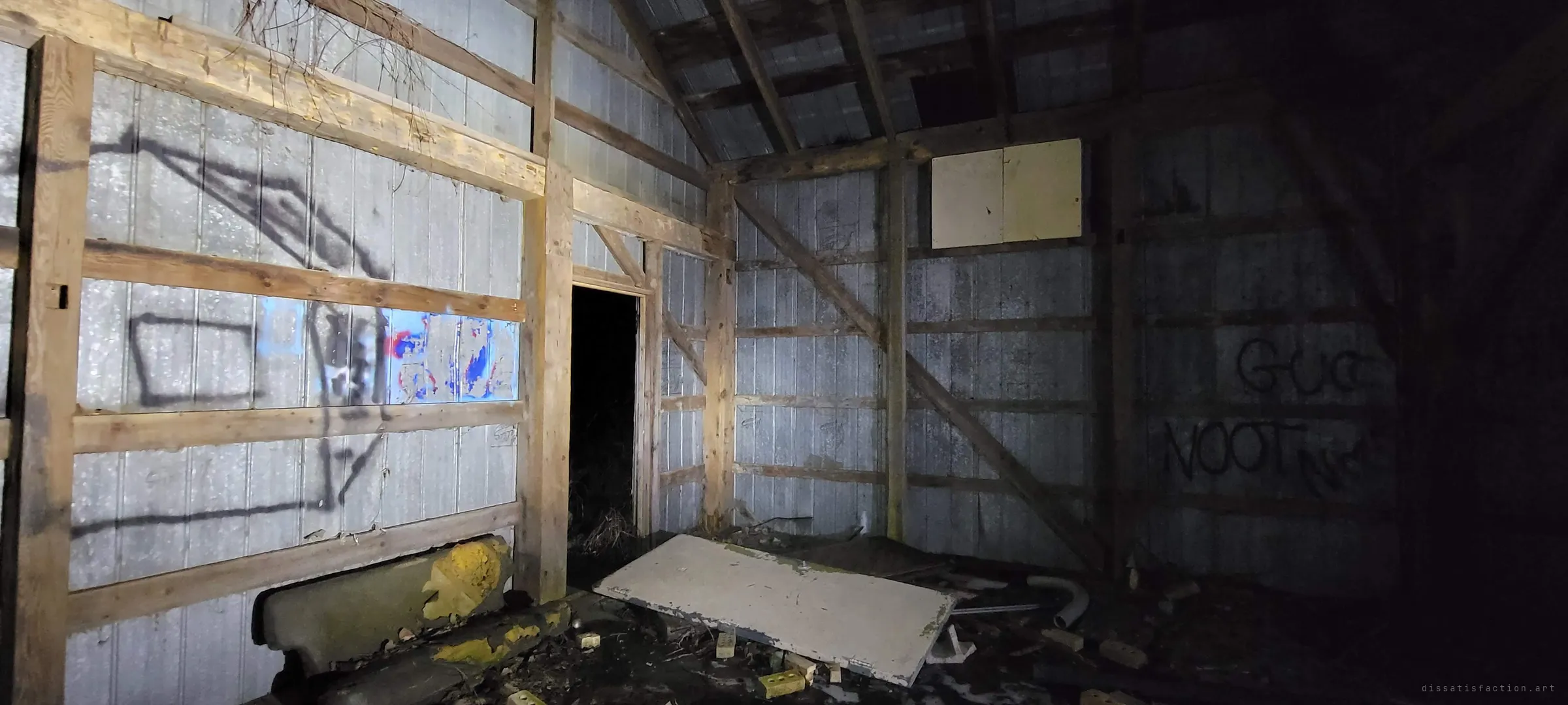 A corner of the barn with pale metal walls, rough timber framing and spray-painted tags, a fallen panel and bricks on the floor. A dark doorway leads out into the night.