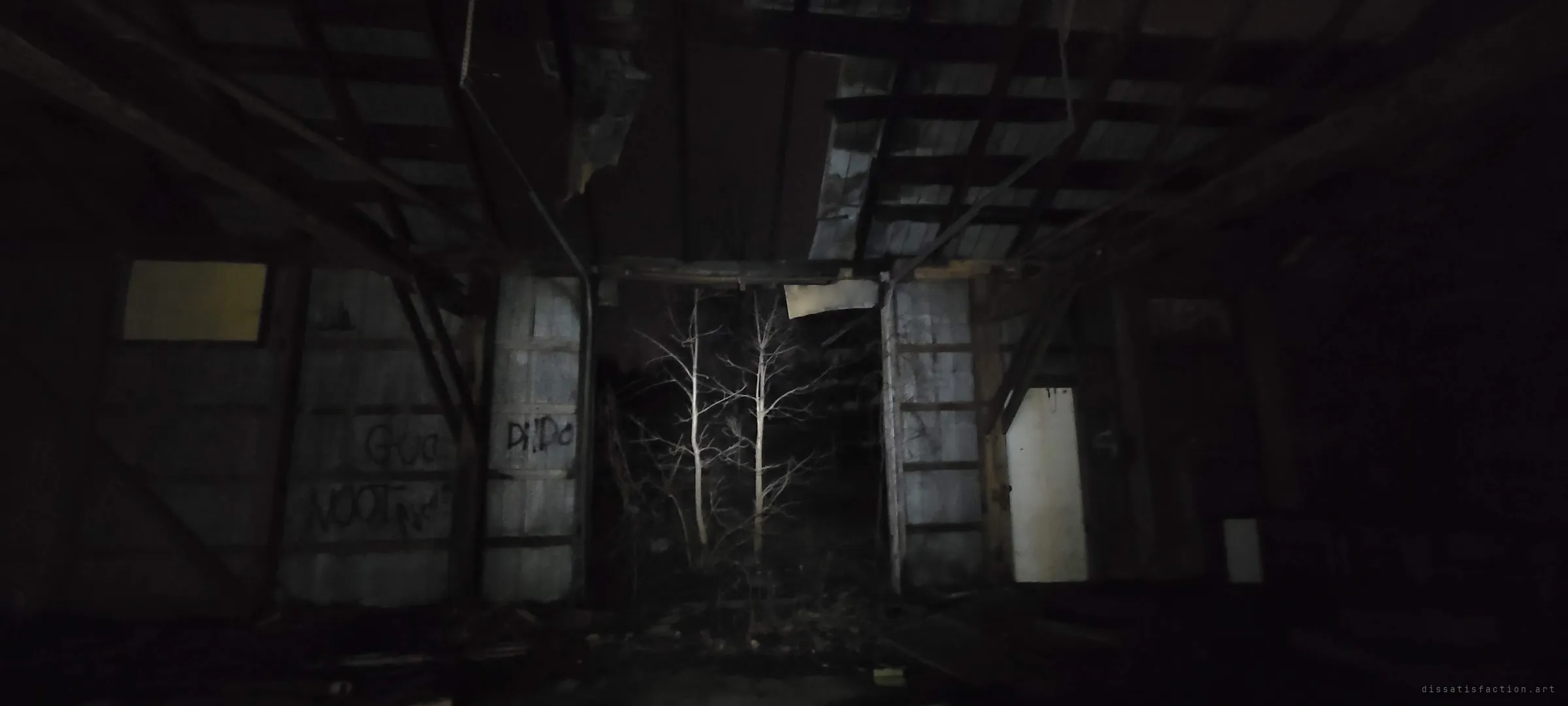 Looking through the barn's missing wall, pale bare trees glow in the torchlight between two tagged metal panels under a broken roof. The woods are already growing into the opening.