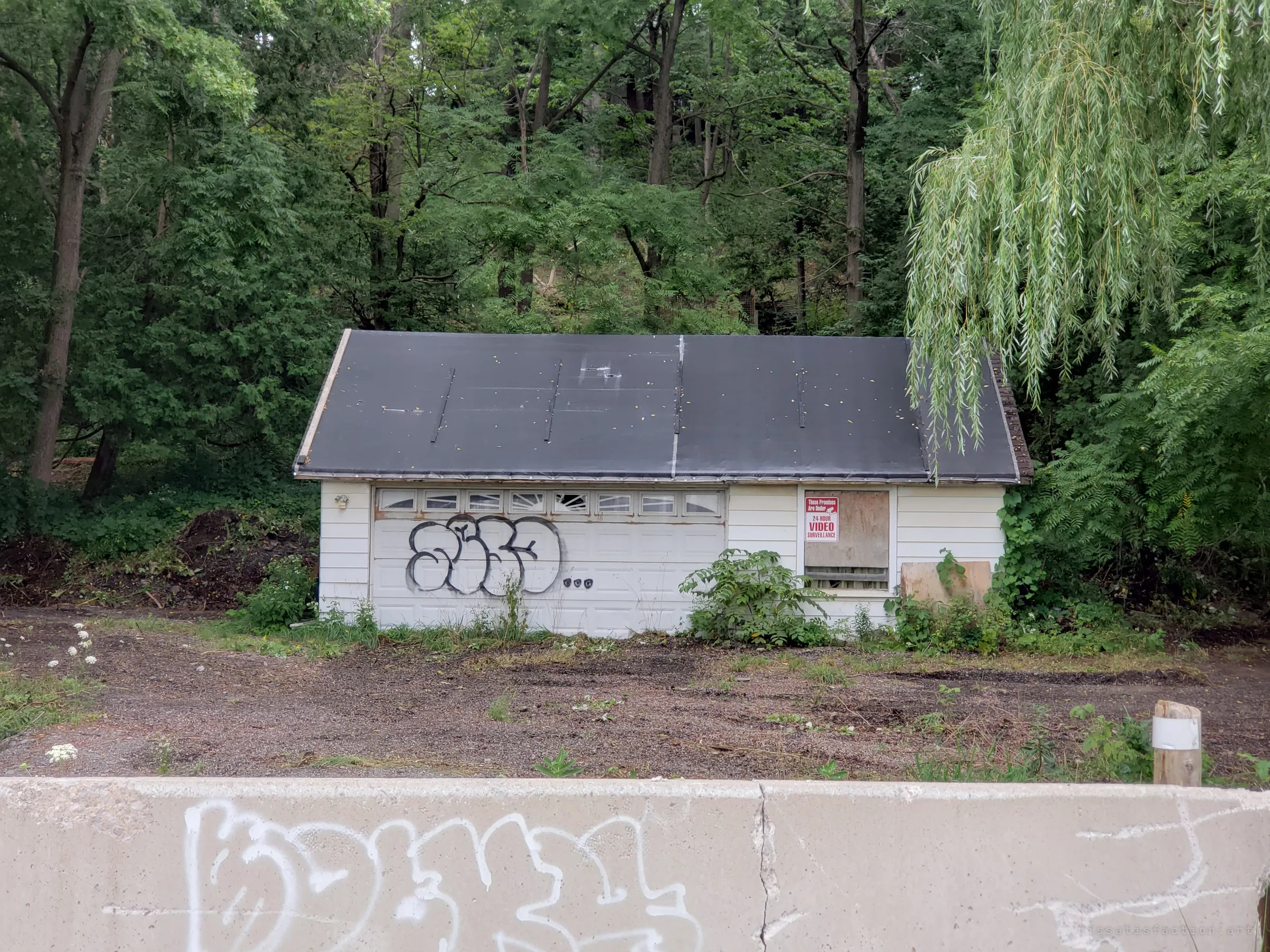 A white detached garage tagged with graffiti, its side window boarded and posted with a generic video-surveillance warning, behind a graffitied concrete barrier. Dense trees and a willow crowd in behind it.