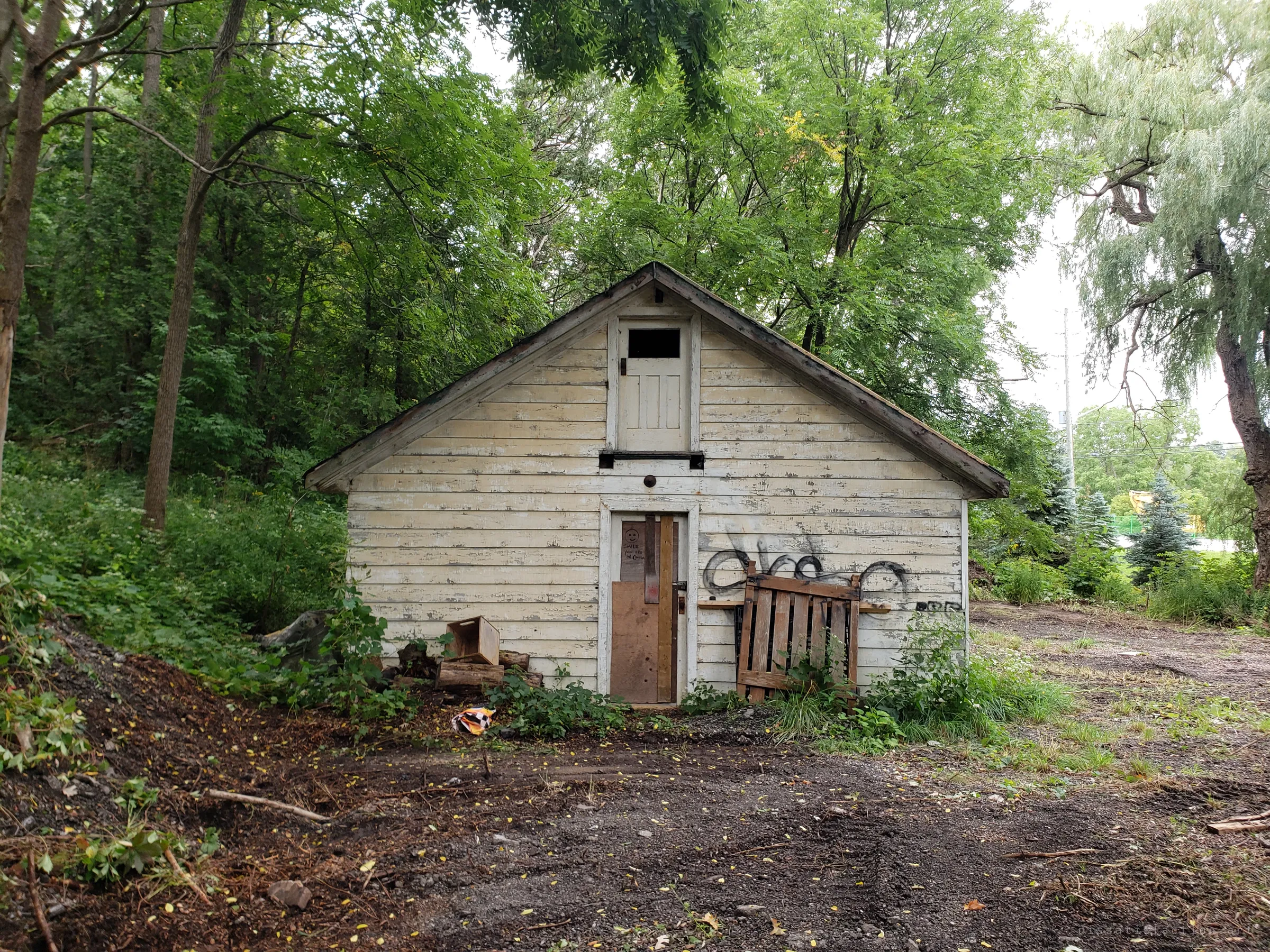 A weathered clapboard outbuilding with a hayloft door, its lower door boarded over and a pallet leaning against a graffitied wall. Freshly scraped ground in front hints at clearing work on the property.