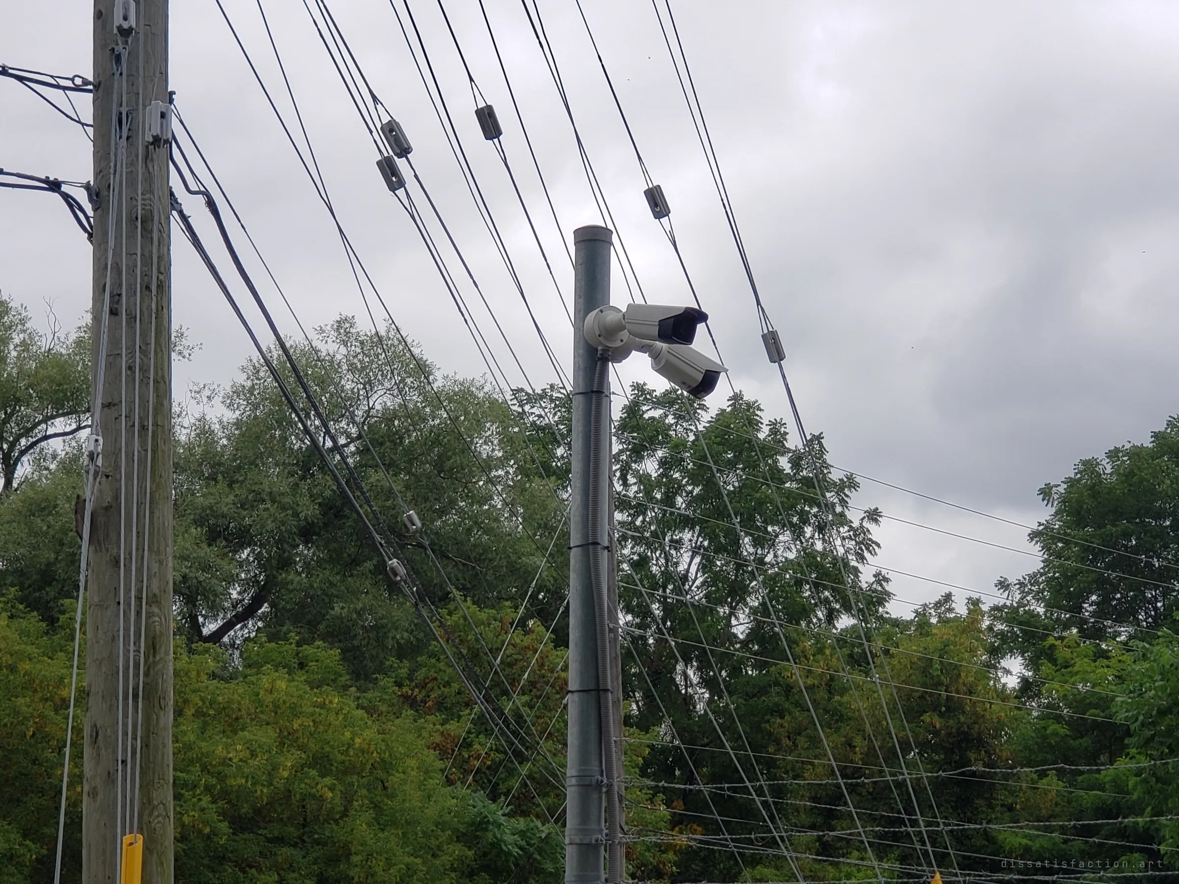 Two security cameras mounted on a steel pole among a tangle of power lines and barbed-wire strands against a grey sky. A reminder that the property was being watched.