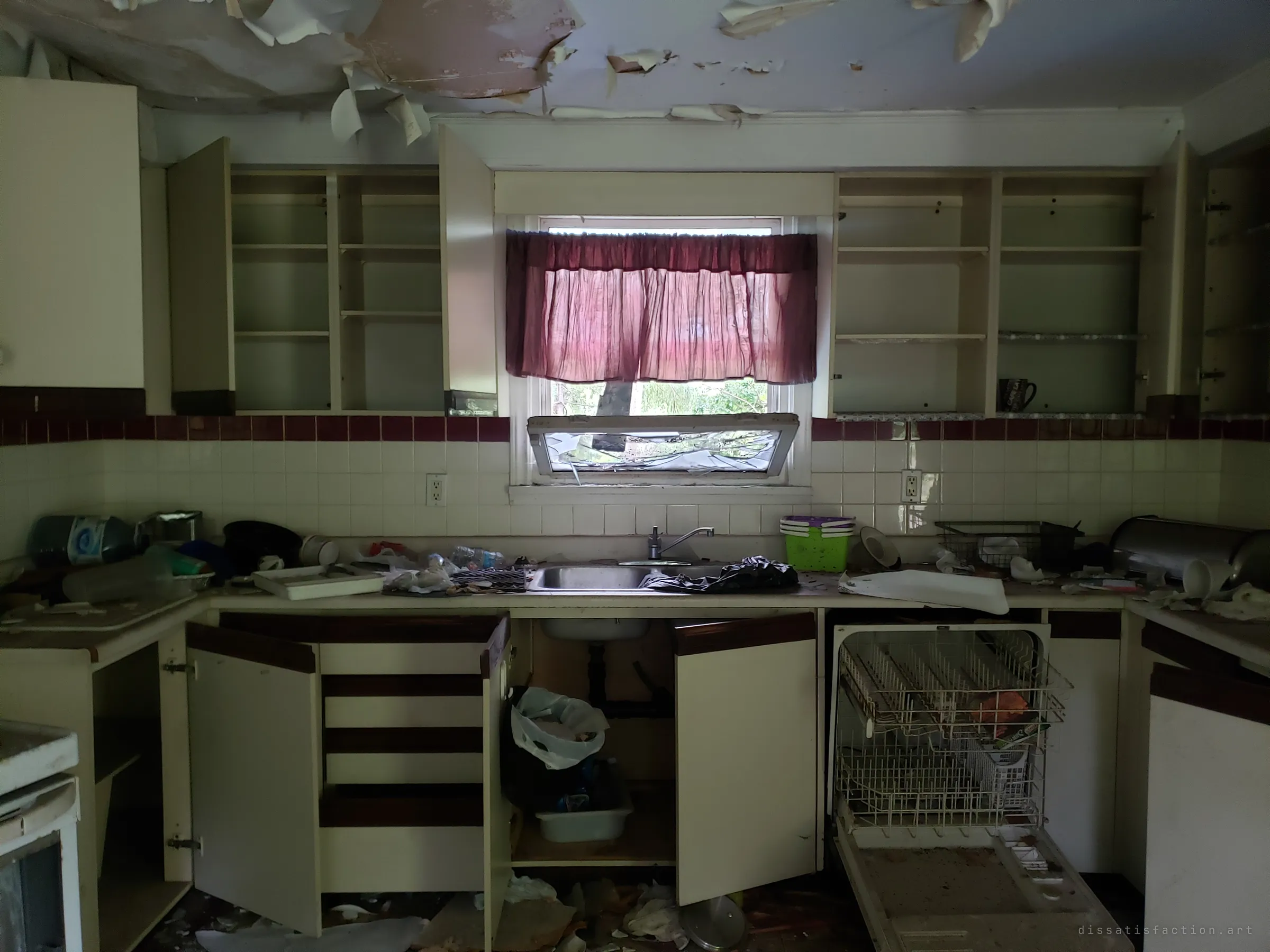 A gutted kitchen with every cabinet door flung open and the shelves empty, the dishwasher hanging open below a smashed window. The ceiling paint peels away in curling sheets above the clutter on the counters.