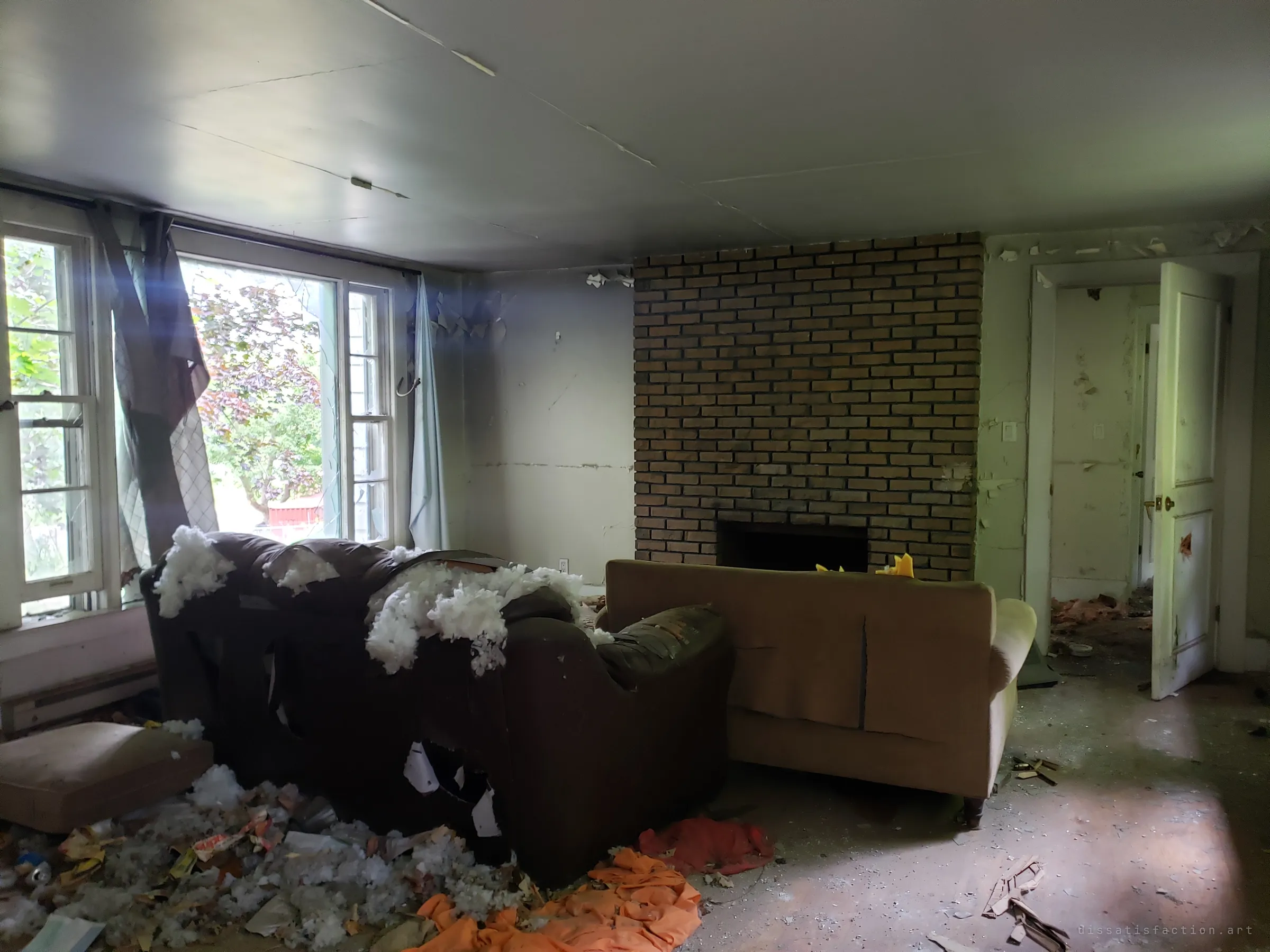 A living room with torn curtains at bright windows, a shredded armchair spilling stuffing and a sofa pushed against a brick fireplace. Litter and fabric cover the floor, and a doorway leads deeper into the house.