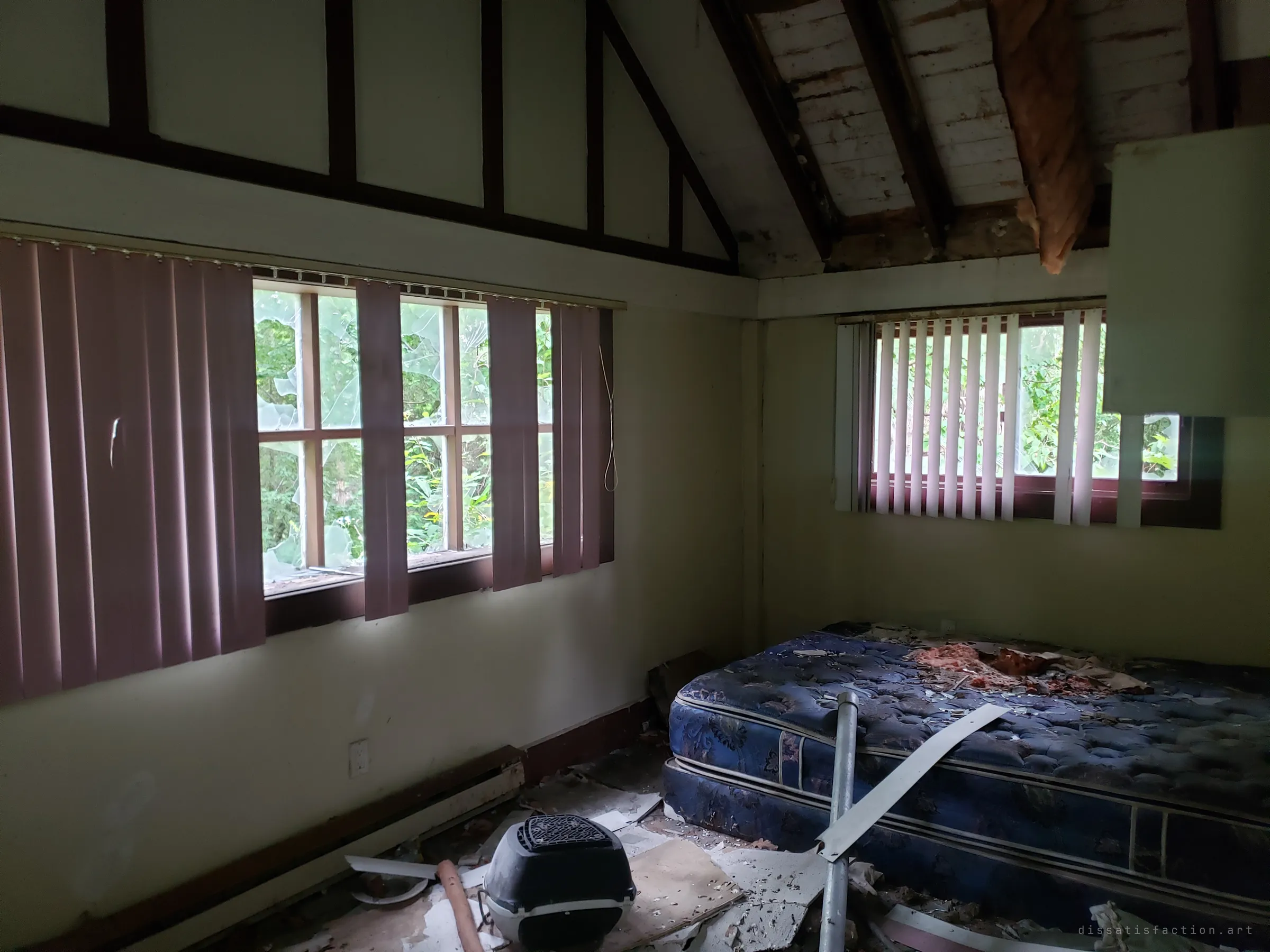 An attic bedroom under a vaulted, open-beamed ceiling, torn insulation hanging above a debris-covered mattress. Faded vertical blinds frame shattered windows with green growth pressing in from outside.