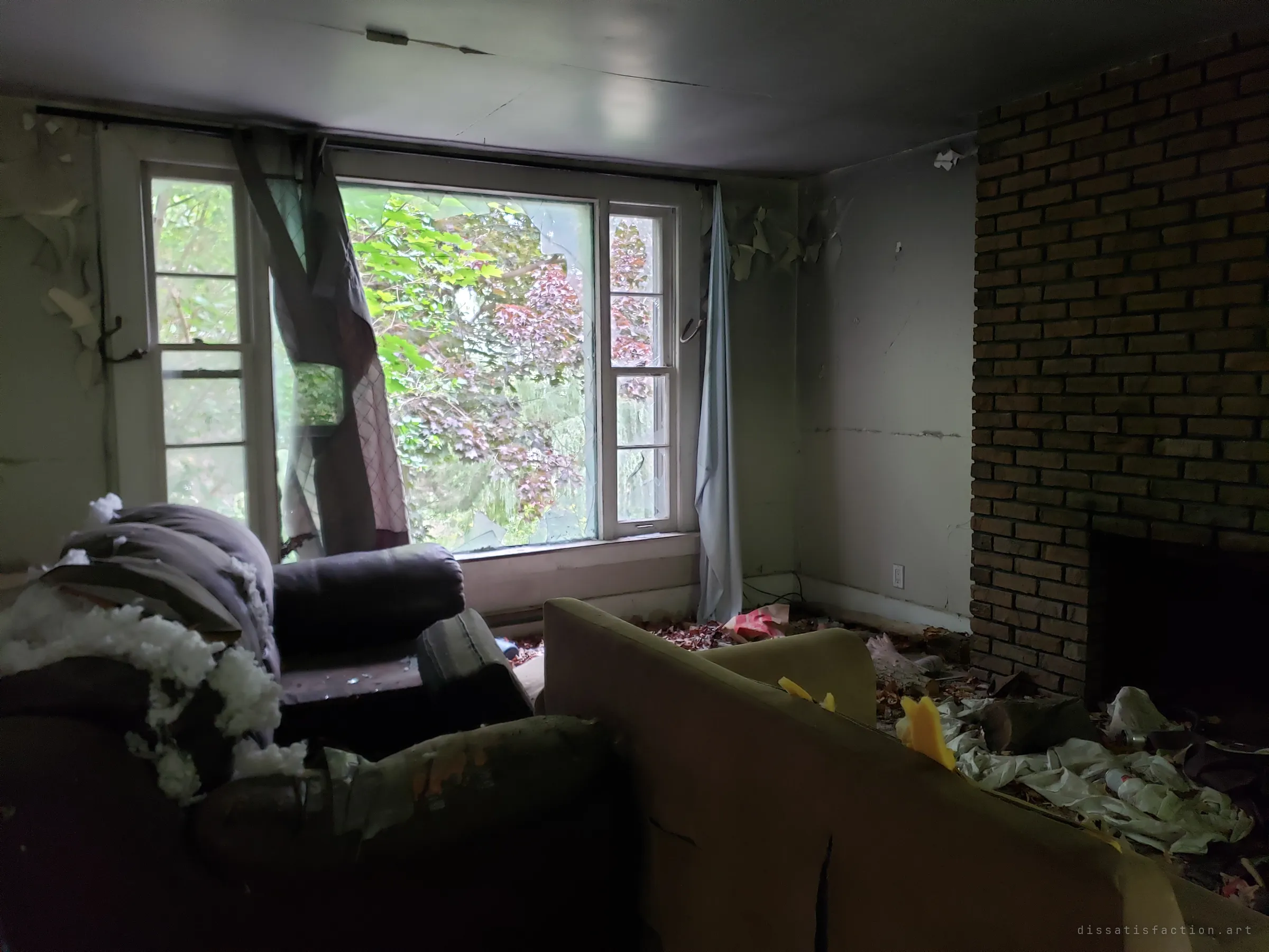 A dim living room with a brick fireplace, torn-open recliners spilling stuffing and a sagging sofa. A curtain hangs through the smashed picture window, where only trees and foliage show outside.