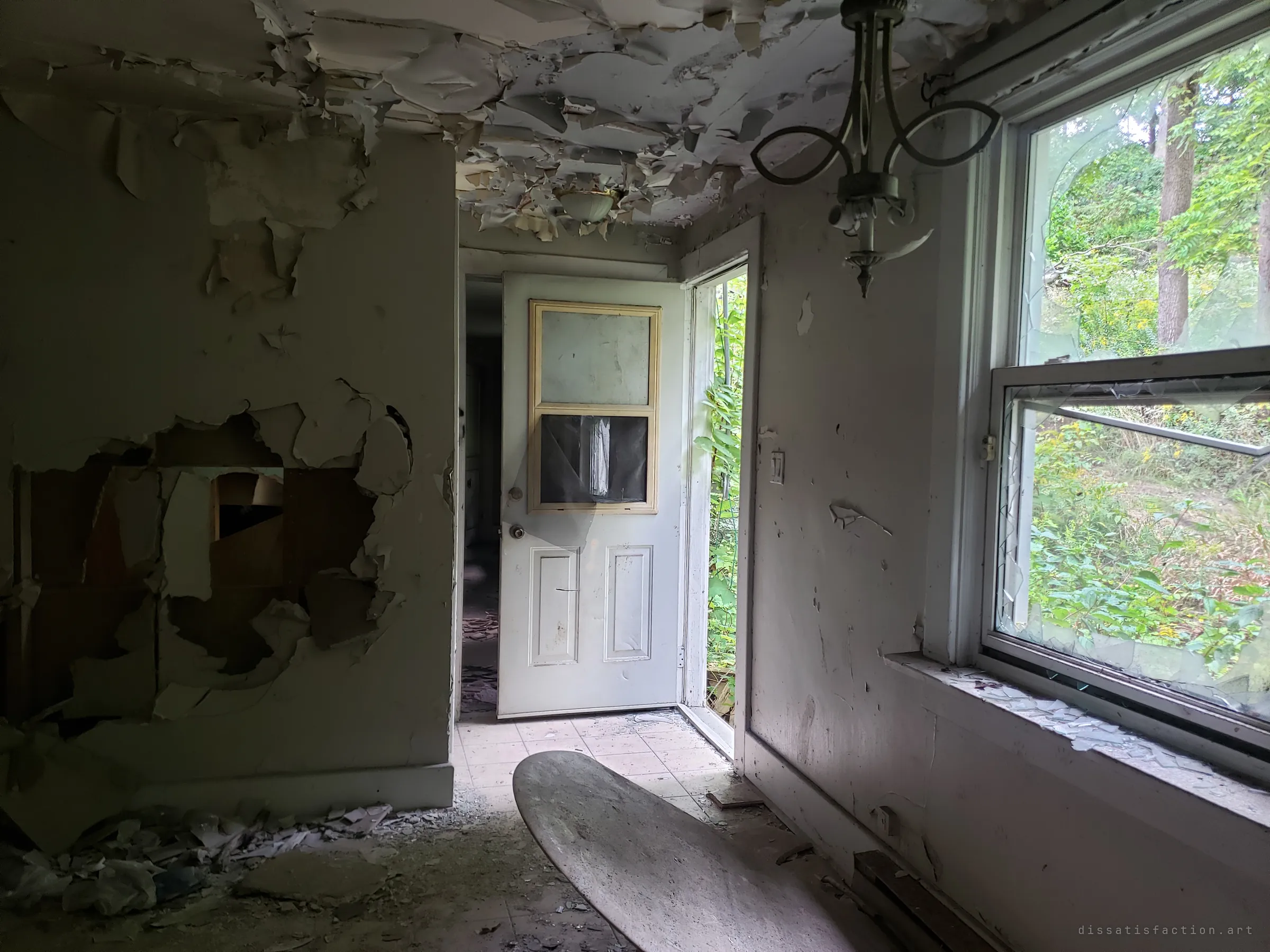 A kitchen of peeling ceiling paint and a gutted wall, a tabletop lying on the tiled floor. The back door stands open to overgrowth and a broken window frames the woods beside the house.