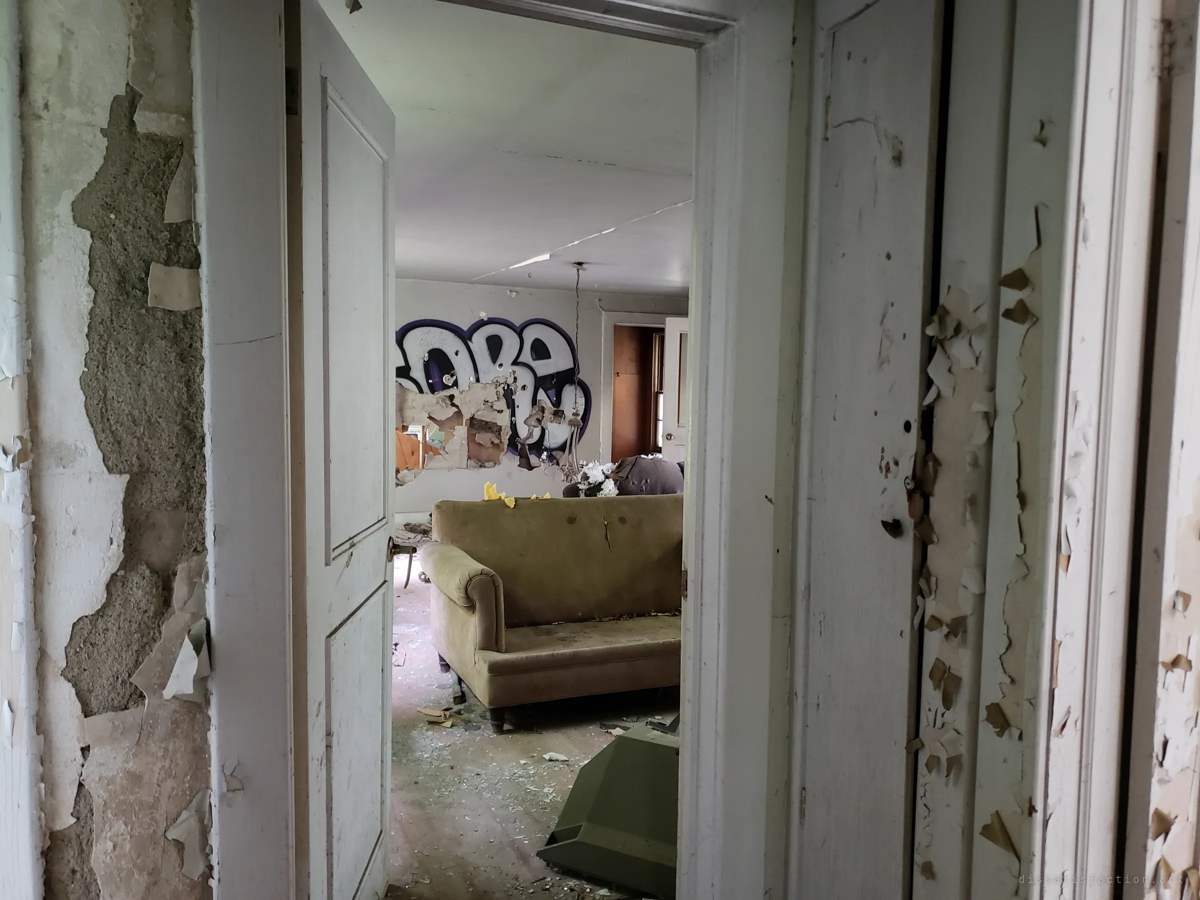 Looking through a doorway of flaking paint into the living room, where a large graffiti piece covers the torn plaster behind a worn olive sofa. The house has clearly been visited and tagged many times.