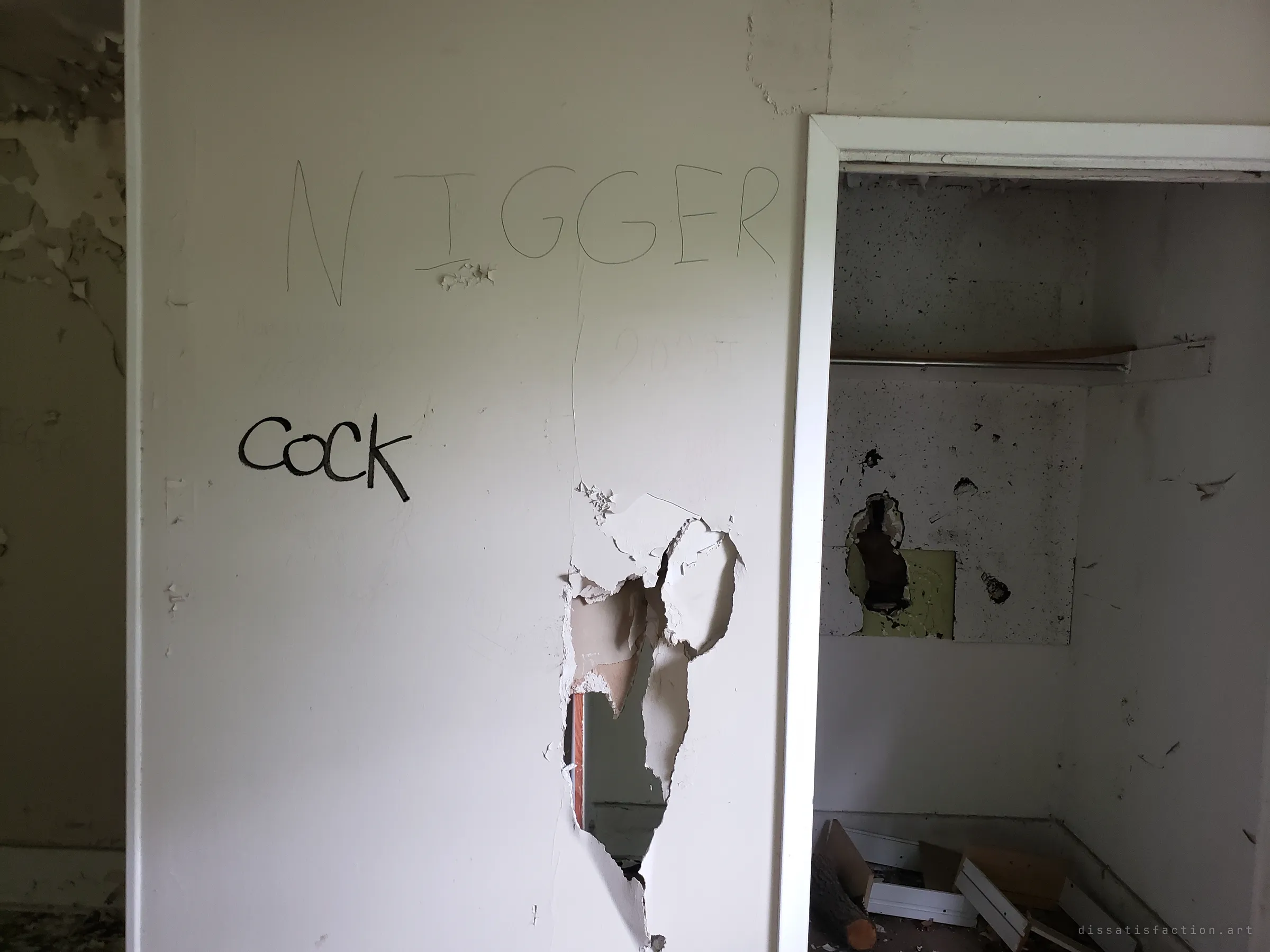 A bedroom wall punched through beside an empty closet, covered in crude pencil and marker graffiti including a slur. Vandals have left their mark on nearly every surface.