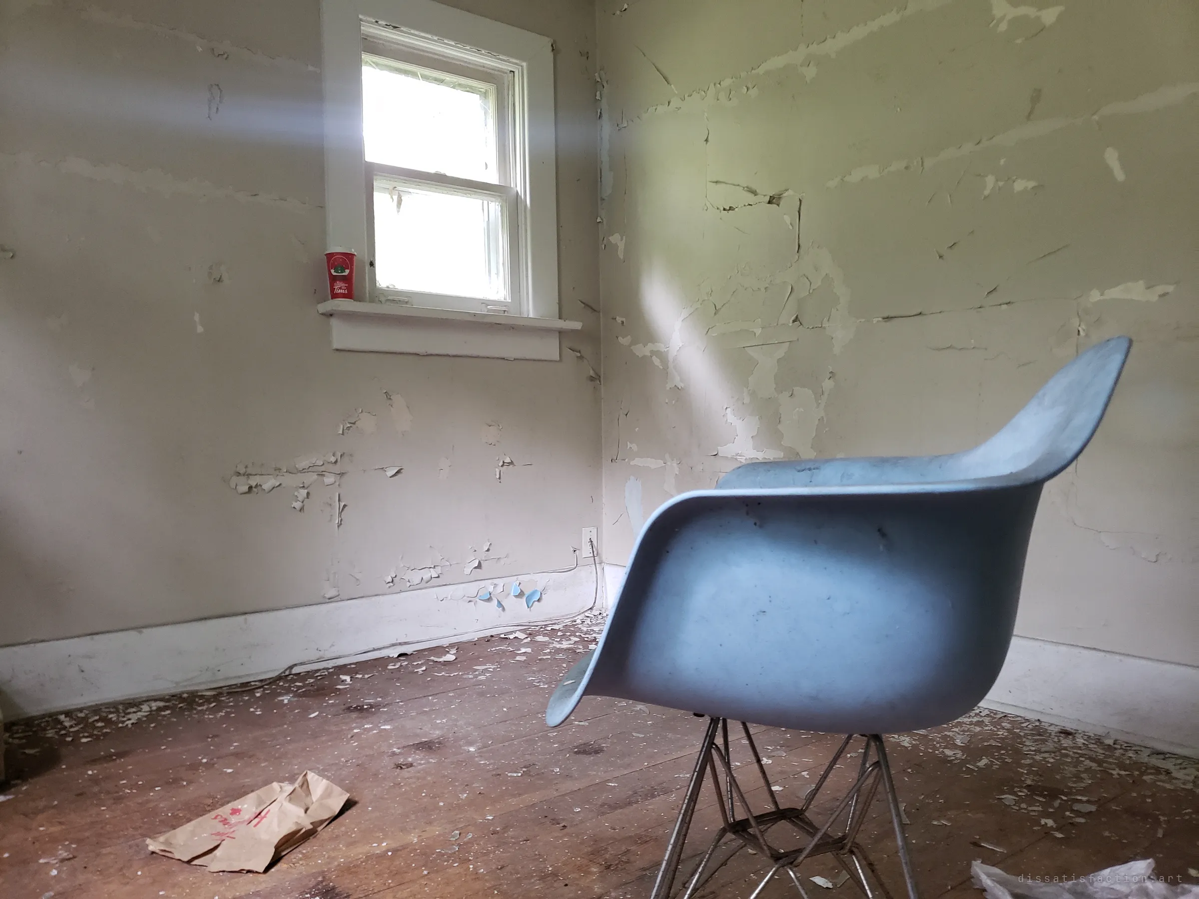 A light-blue shell armchair sits alone in a small room of peeling paint, a shaft of light falling across the wall. A takeaway coffee cup left on the windowsill hints at recent visitors.