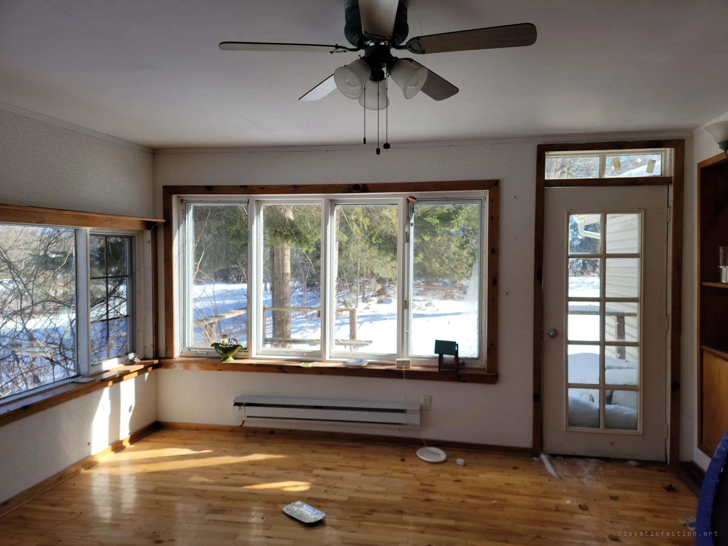 A sunroom with pine-trimmed windows looking out onto snow and evergreens, a ceiling fan overhead and bright winter light across the wood floor. The house still feels lived-in, just emptied of people.