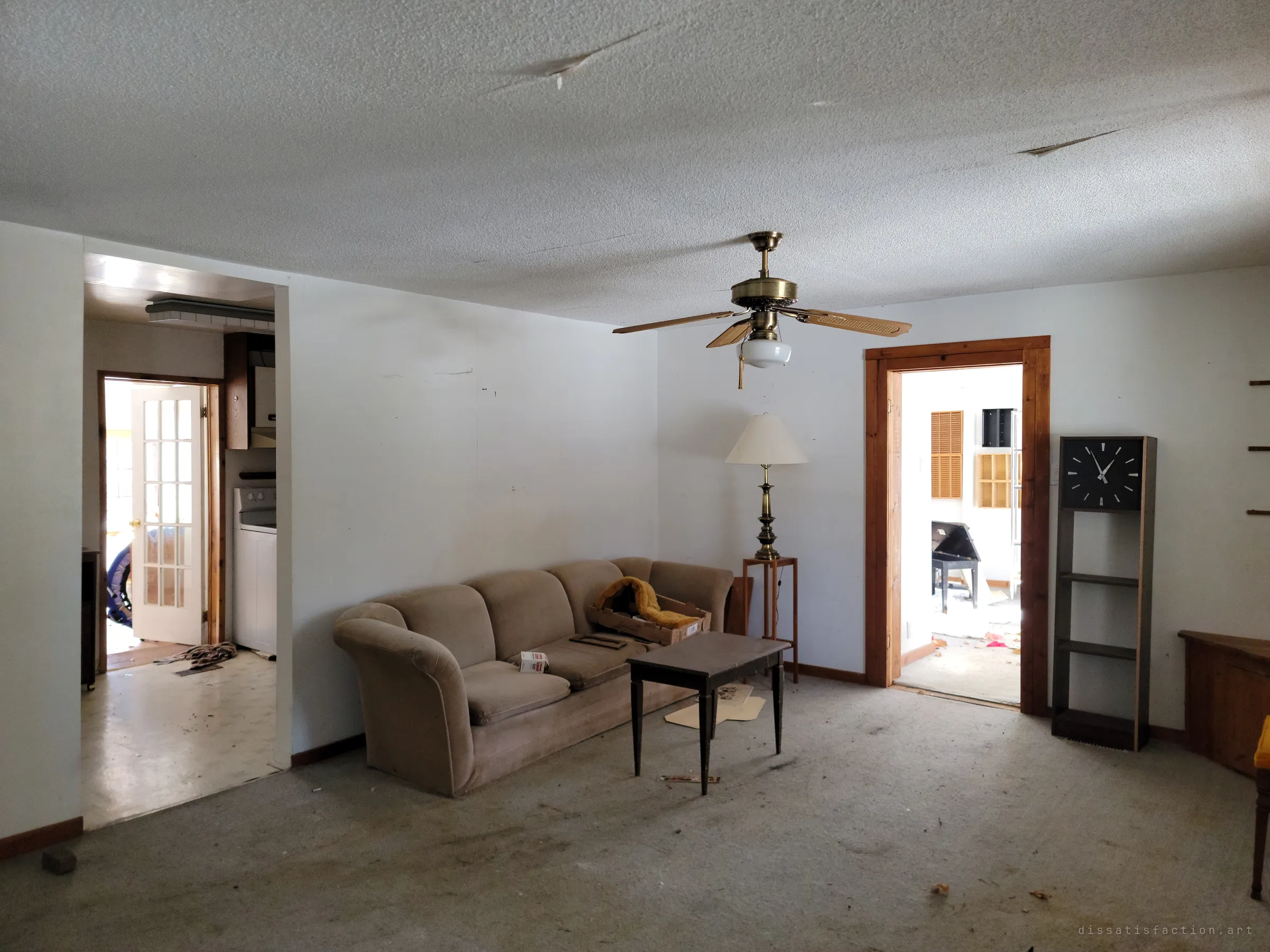 A carpeted living room with a worn beige sofa, a brass floor lamp and a tall clock shelf, with doorways leading to the kitchen and a bright back room. The furniture was left where it stood.