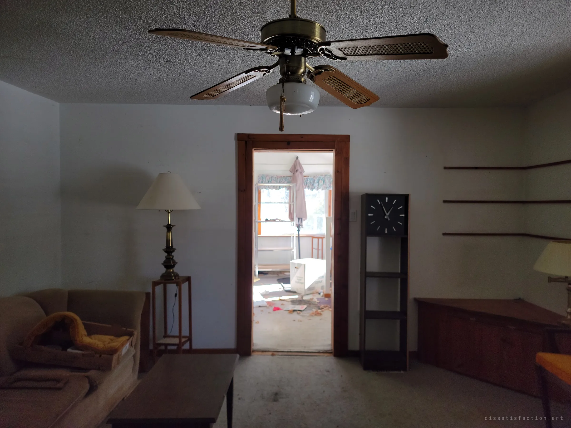 Looking under a brass ceiling fan through a pine-framed doorway into a sunlit room strewn with debris and a folded patio umbrella. The dim living room frames the mess beyond.