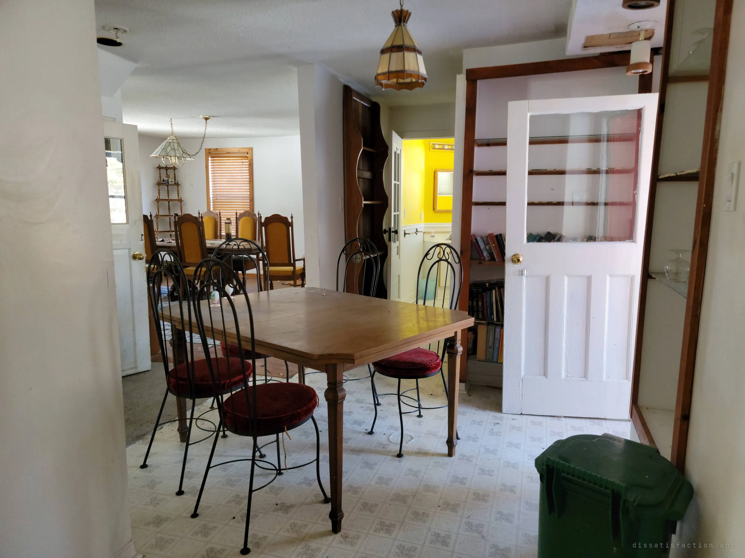 A kitchen dining nook with a wooden table and wrought-iron chairs on red velvet cushions, a bookshelf and a yellow bathroom glimpsed past an open door, and a formal dining room beyond. Every room still holds its furniture.