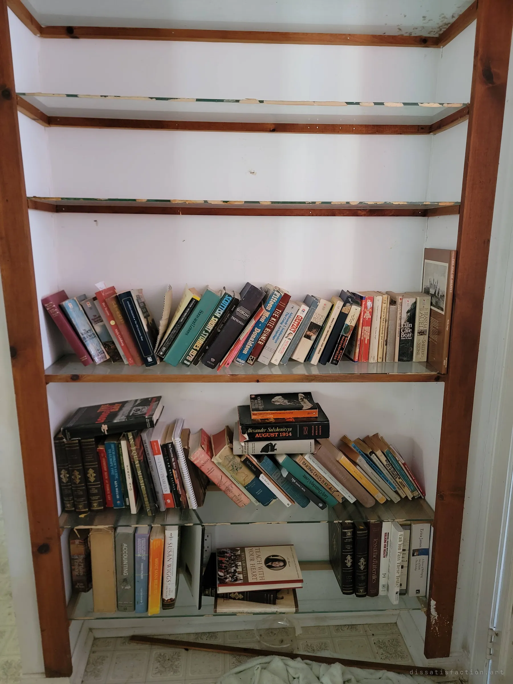 A built-in shelf with its upper glass shelves empty and its lower ones crowded with paperbacks, cookbooks and hardcovers. The reading left behind hints at who lived here.