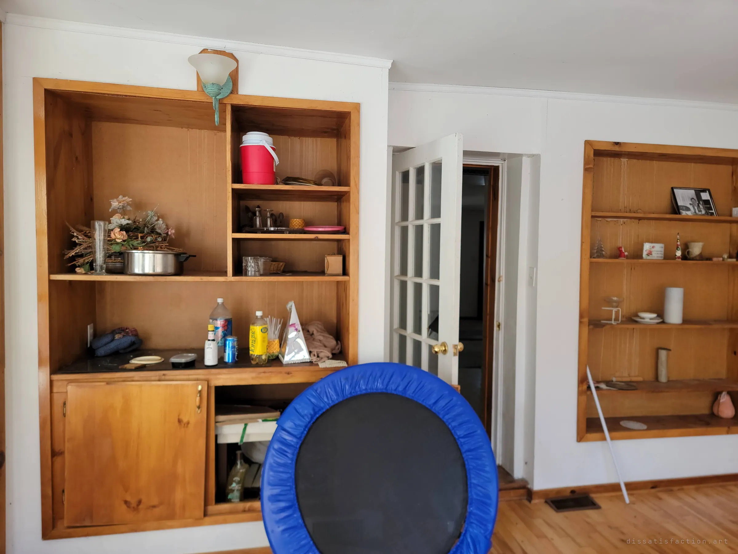 Pine built-in cabinets holding a red cooler, silk flowers, pots and half-finished drinks, with a blue mini trampoline in the foreground and the photo niche on the far wall. Everyday clutter frozen mid-use.