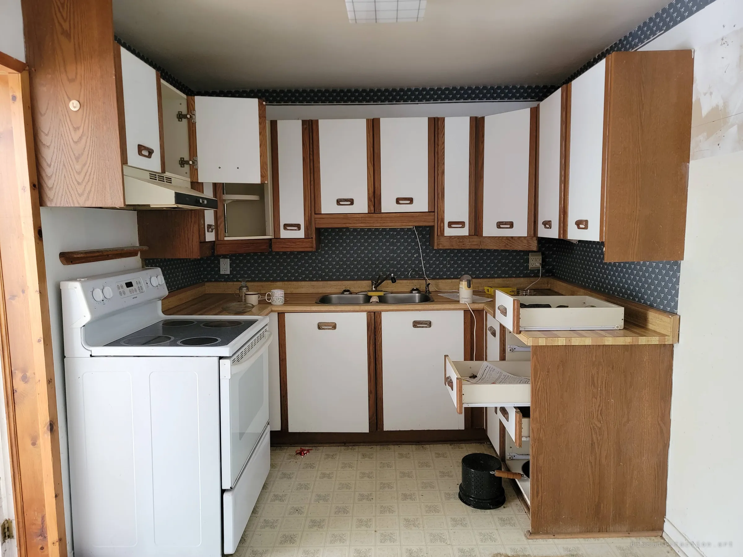 A U-shaped kitchen with white-and-oak cabinets, several doors hanging open and drawers pulled out, a stove and a pot left on the floor. Patterned navy wallpaper frames the counters, the room abandoned mid-clear-out.