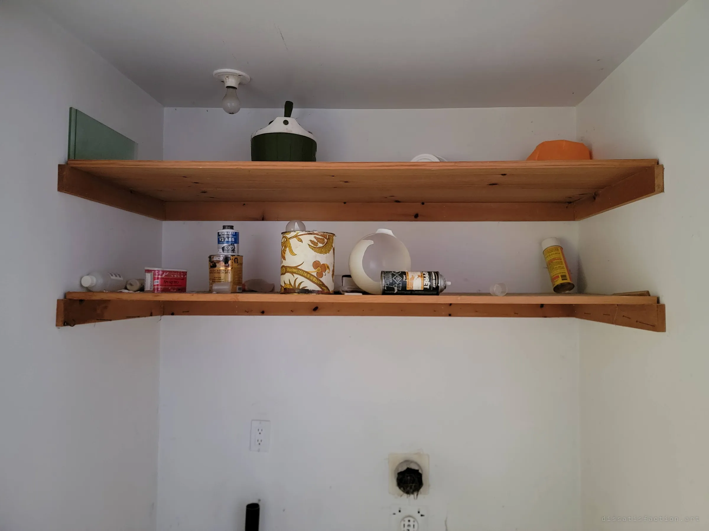 Two pine shelves in a bare white laundry nook holding old paint tins, spray cans and a vintage canister, with an unplugged dryer outlet below. A quiet still life of a utility room nobody uses any more.