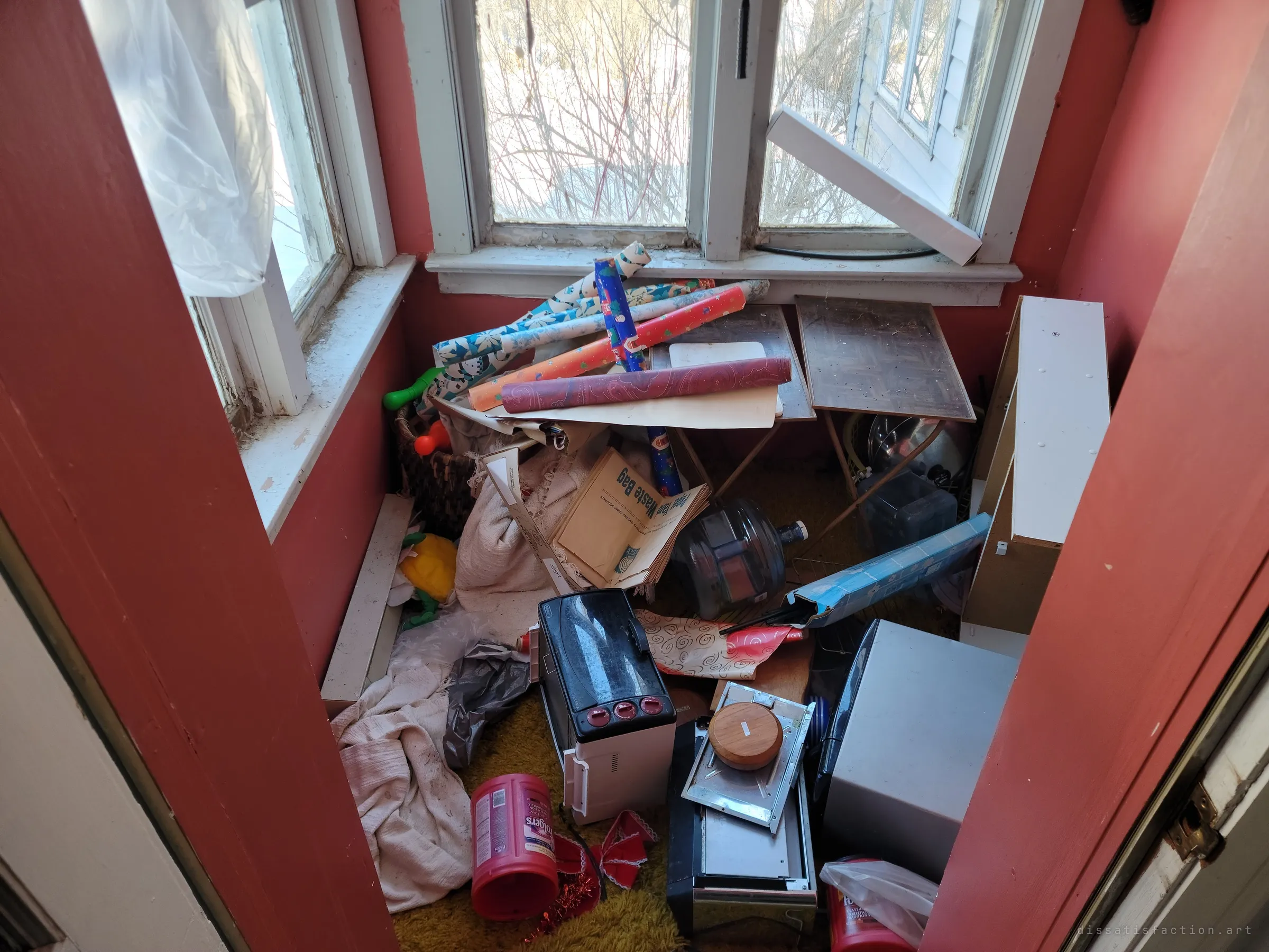 A small red-walled porch piled with wrapping-paper rolls, a toaster oven, folding tables and cast-off household clutter under a bright winter window. The room has become a dumping ground for whatever was left.