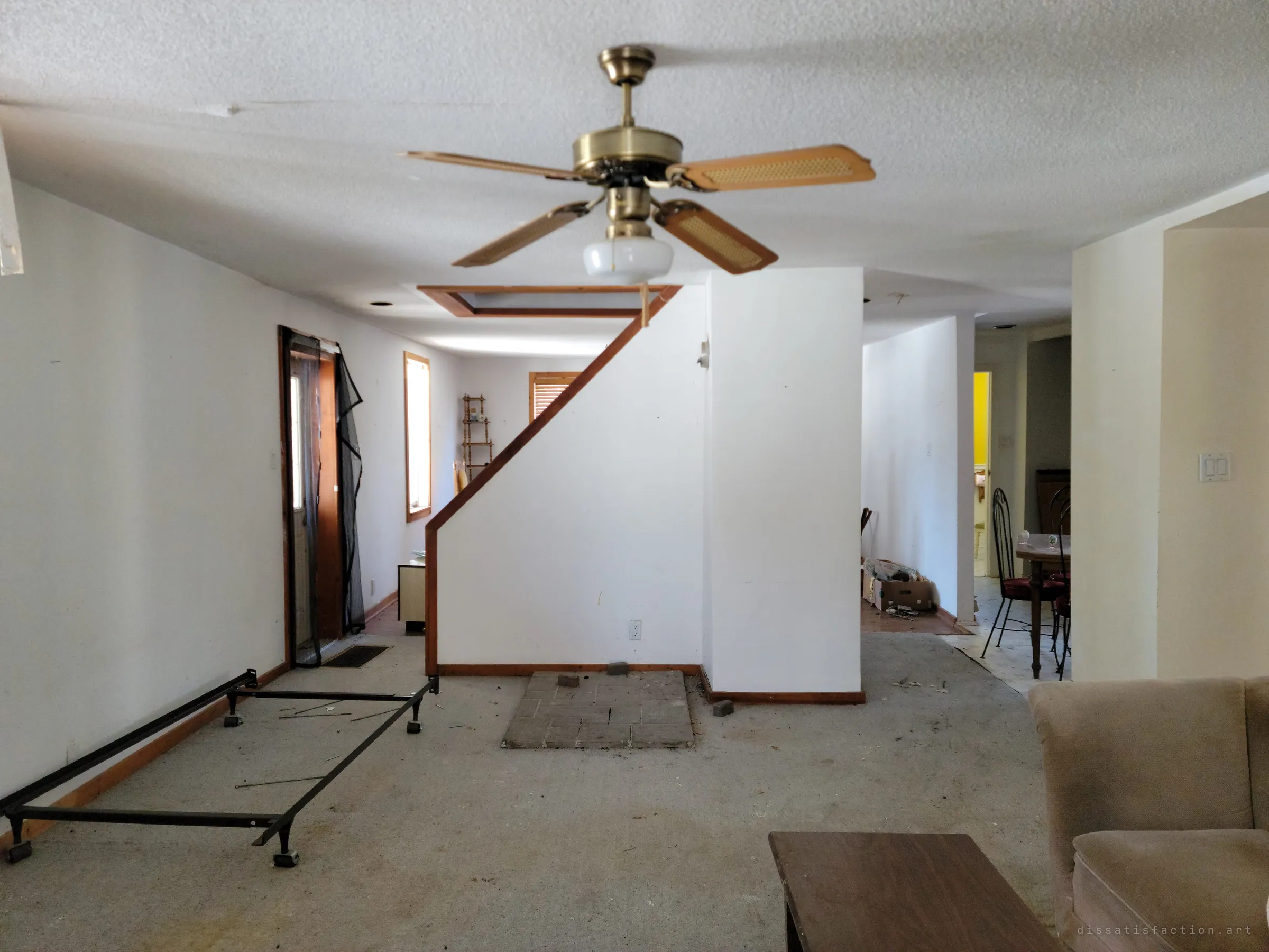 A stripped open-plan living room with a brass ceiling fan, a bare bed frame on the grey carpet, the hearth pad of a removed stove and a staircase wall leading toward the dining area. The house is emptied but still holds its layout.