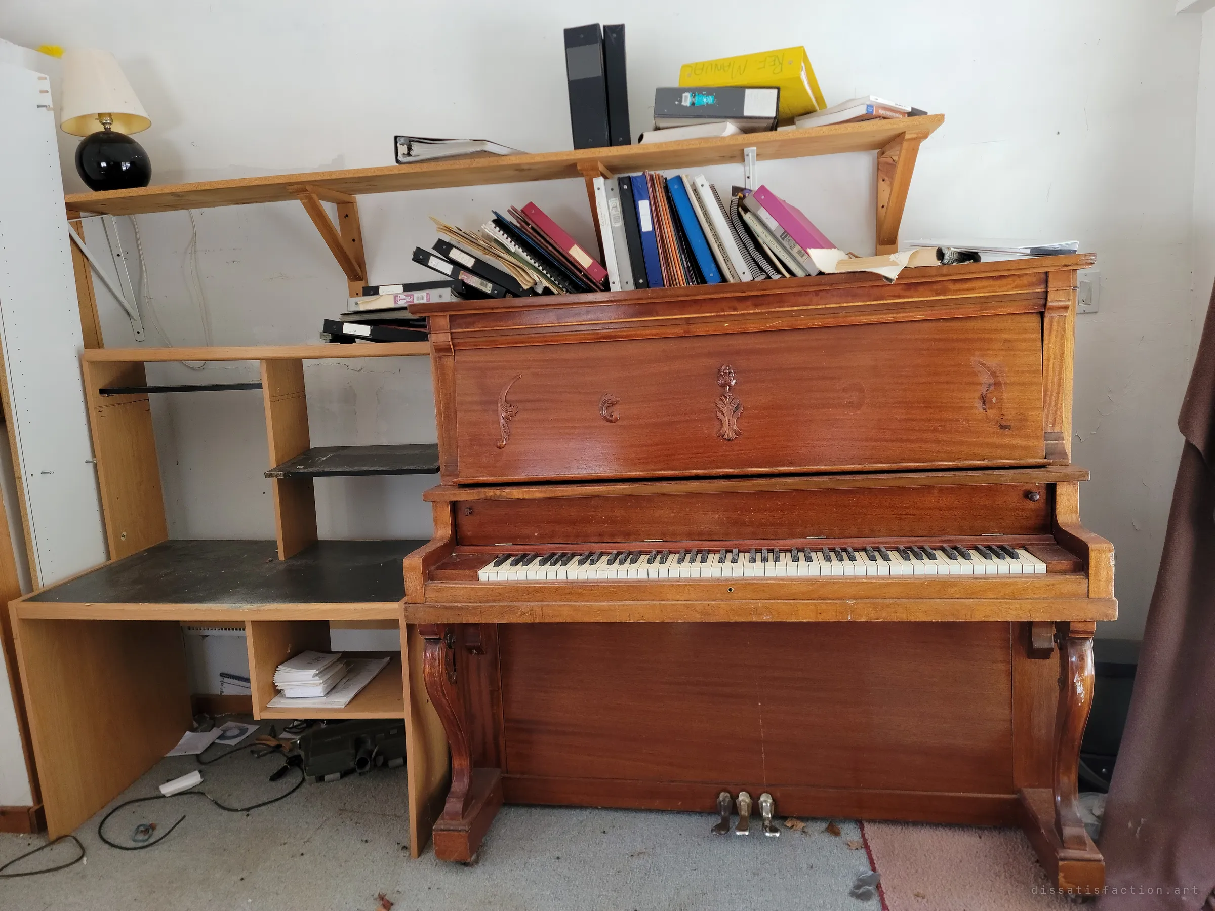 An old upright piano with carved panels, its top crammed with music binders and songbooks, beside an empty desk unit and a black ball lamp. The music stayed when the people didn't.