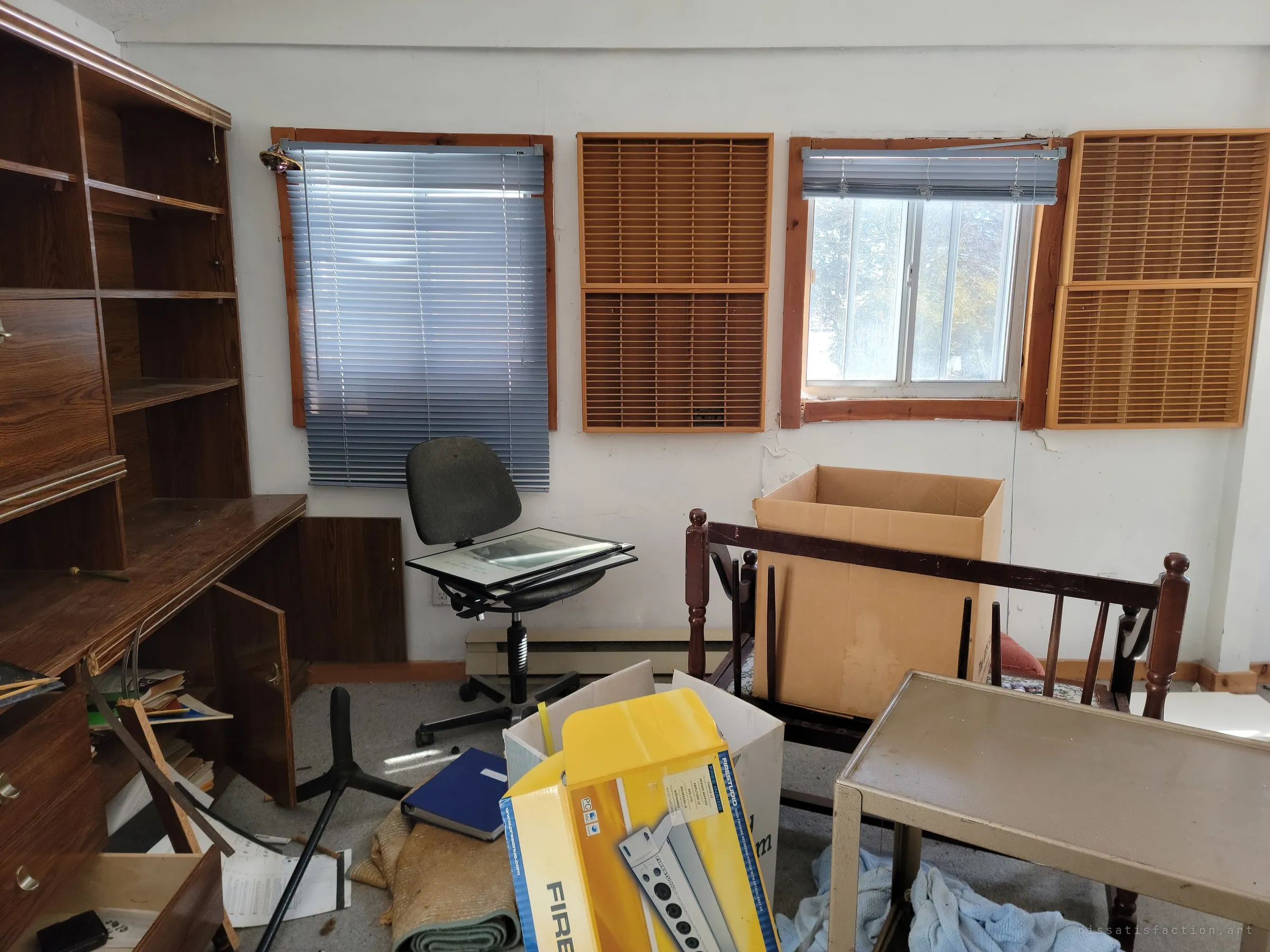 A cluttered home office with wall-mounted cassette racks between the windows, a tipped office chair holding framed prints, and an audio-interface box among scattered papers. Someone recorded or collected music here.