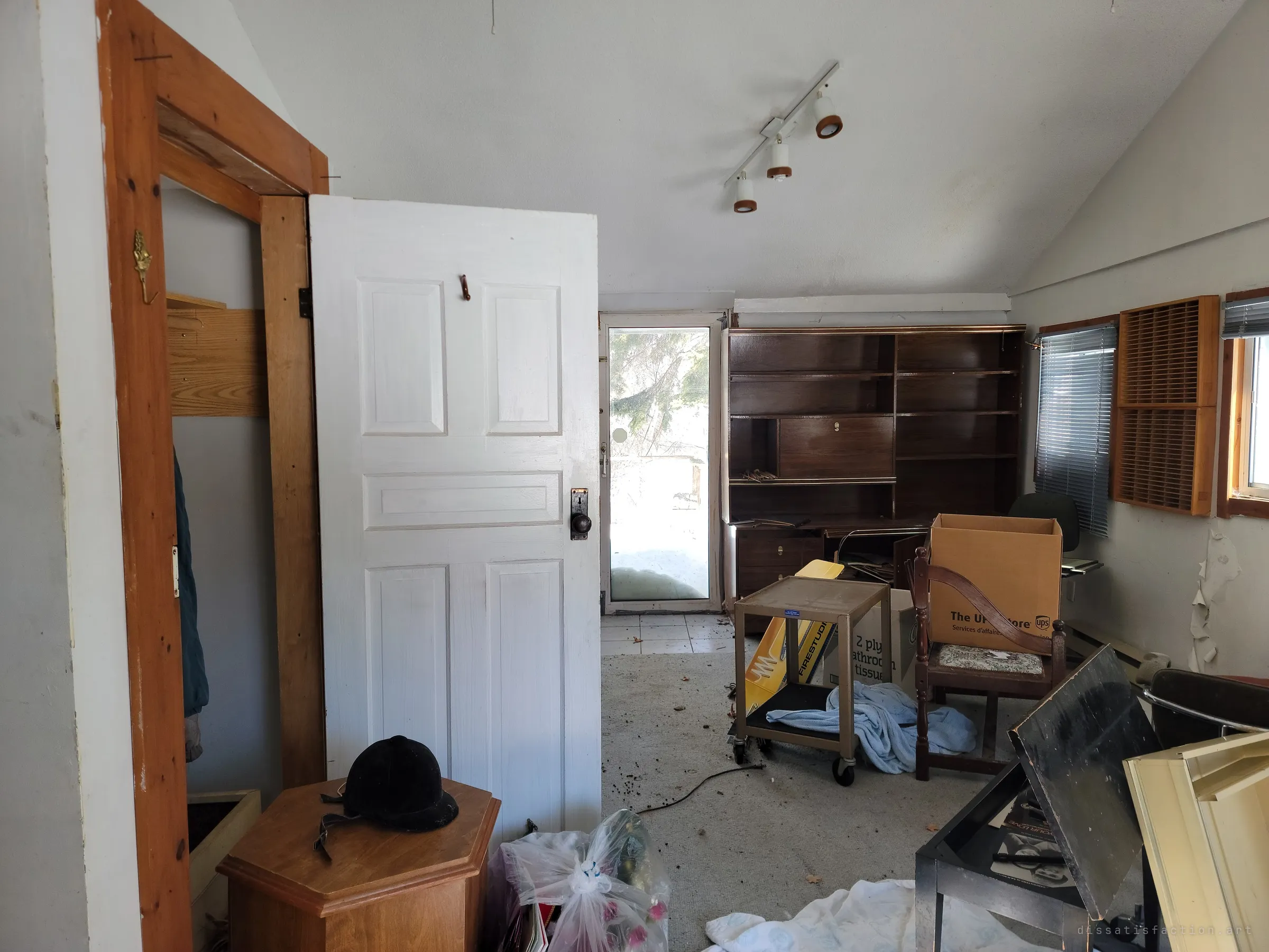 A vaulted back room seen past an open panelled door, with a dark wall unit, track lights, moving boxes and a riding helmet left on a side table. Snow glare pours through the glass door at the far end.